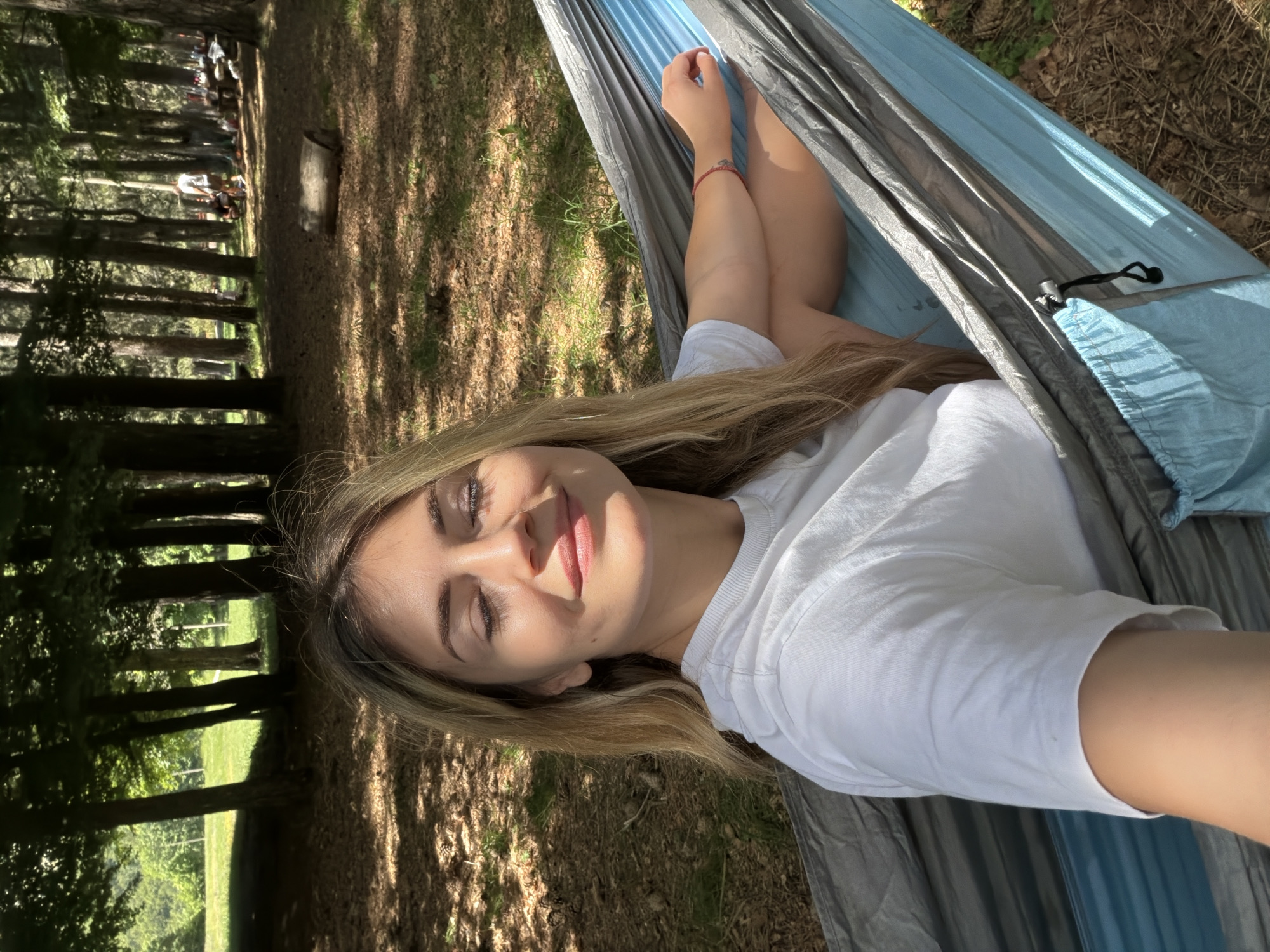 A fair-skinned woman with long, light brown hair is positioned in a two-toned grey and light blue fabric hammock, occupying the central and lower-left portions of the image. Her eyes are closed, and she exhibits a gentle smile, with her face illuminated by direct sunlight. She is wearing a plain white short-sleeved t-shirt. Her left arm, adorned with a red string bracelet on her wrist, is bent and resting on the hammock's material. A black drawstring or cord is visible on the lower right edge of the hammock.
The background consists of a dense arrangement of tall, slender trees with green foliage, creating an overhead canopy that filters sunlight onto the ground. The ground is covered with dry leaves, pine needles, and sporadic patches of green grass, showing distinct patterns of dappled sunlight and shadow. Several pine cones are visible on the ground near the woman's head. In the mid-right background, a small, cut log rests horizontally on the ground. Further in the background, among the trees, several blurry figures of other individuals are discernible, along with what appear to be outdoor seating or picnic areas, indicating a recreational environment. This scene is located in Mali Bostur, Montenegro.