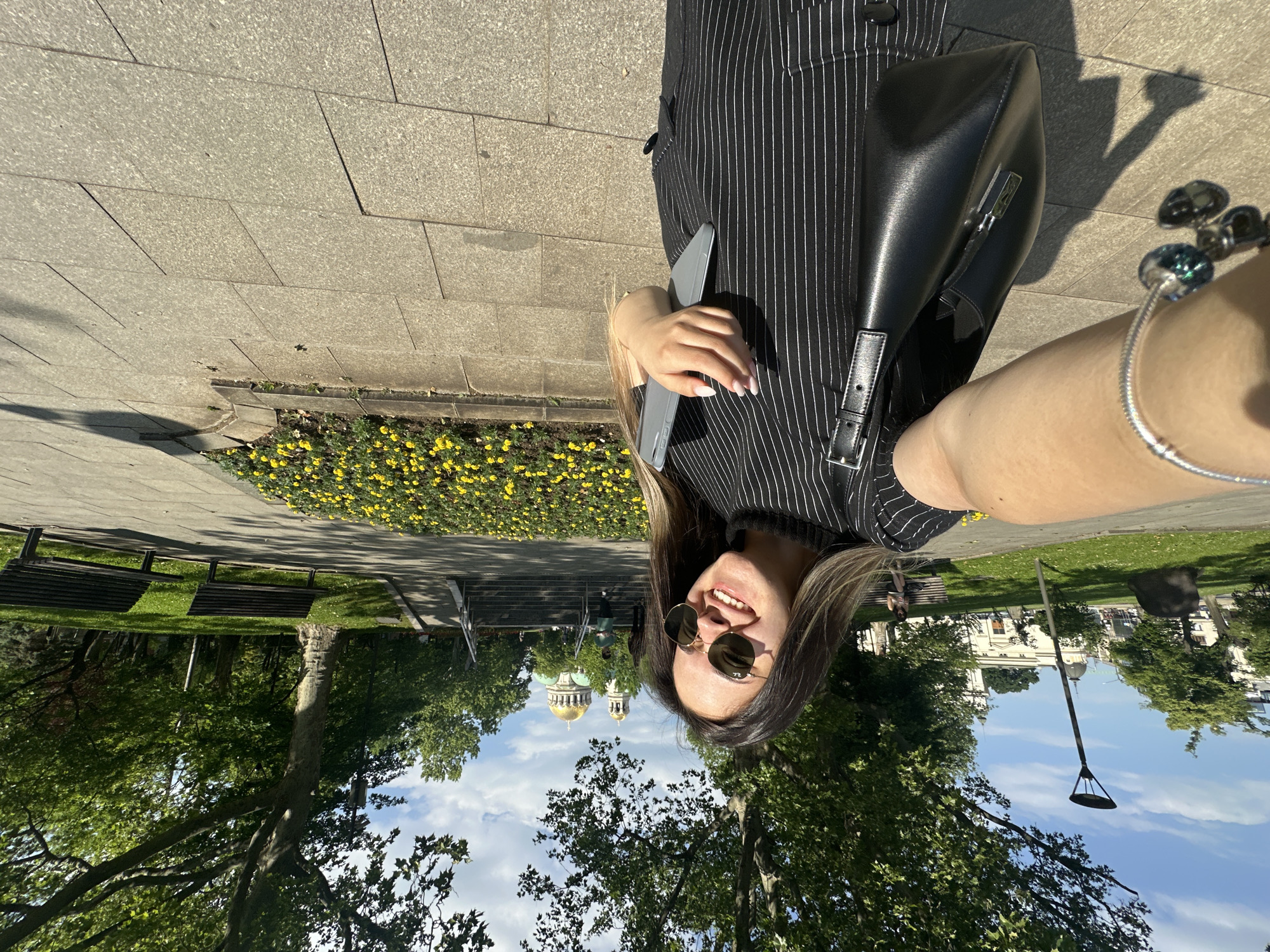 The image shows a young woman posing for a selfie outdoors in Sofia, Bulgaria. She has brown hair, is wearing sunglasses, and a striped black and white outfit with a black purse. She is holding a tablet in her hand. The background includes trees, a green lawn, and a building with a distinctive golden dome, identified as the Alexander Nevsky Cathedral, placing the location in the city center of Sofia. A flowerbed with yellow flowers is also visible. A streetlight stands among the trees. The sky is partly cloudy.