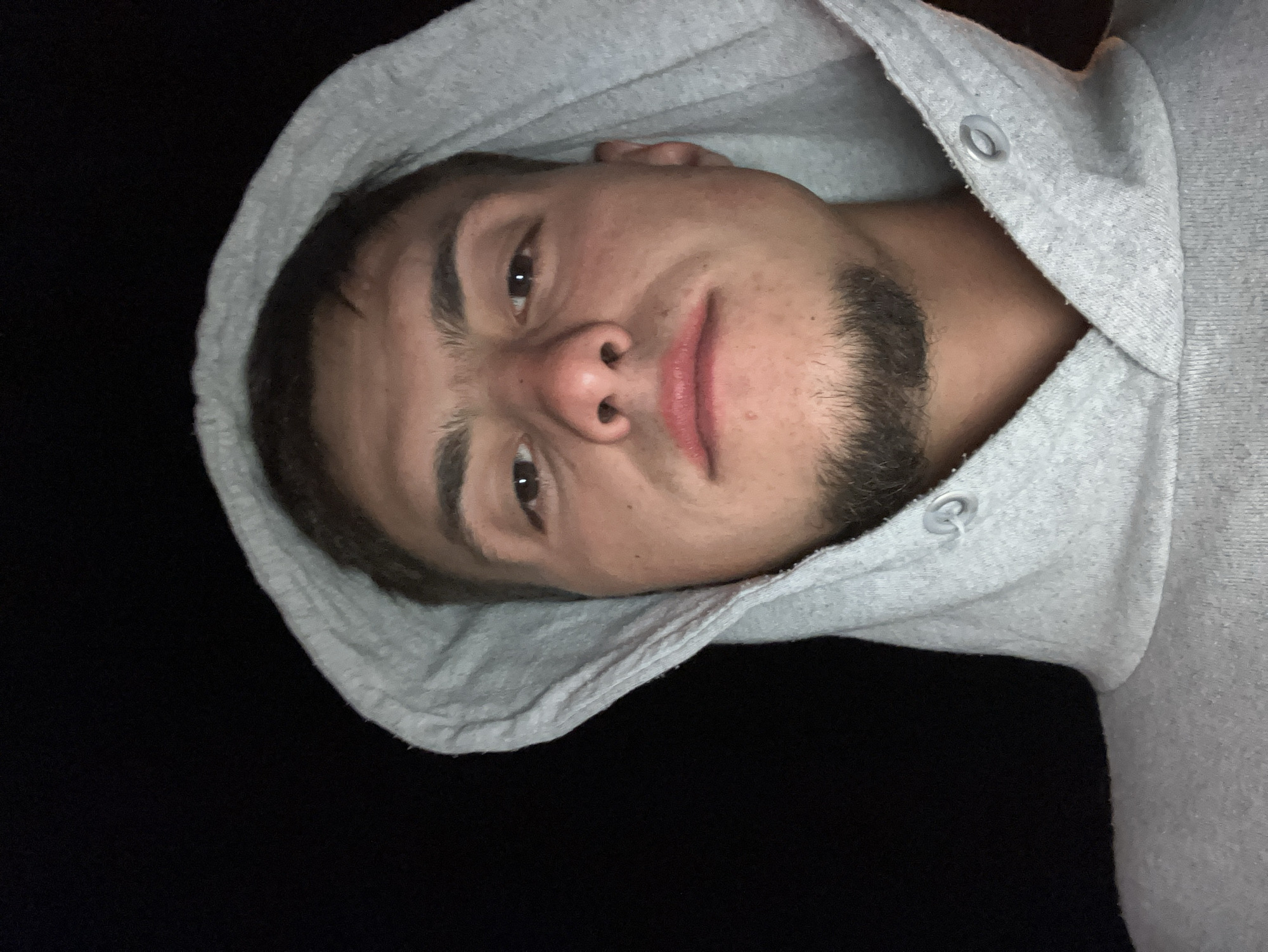 A young man with brown hair and a short beard is wearing a grey hoodie. The hood is up, partially framing his face. He is looking directly at the camera with a slightly pursed expression. The background is dark. There is no evidence to determine the location as Krastevich, Bulgaria.