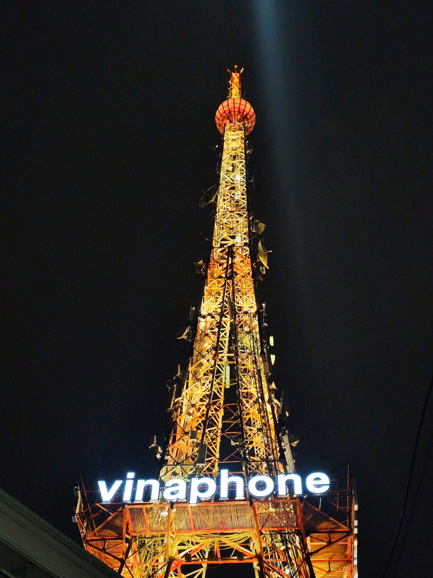 A low-angle, nighttime shot of the Vinaphone tower in Da Lat, Vietnam, showing the illuminated metal structure against a dark sky. The tower has a lattice-like construction, with numerous antennas attached. The word "vinaphone" is spelled out in large, illuminated white letters near the base. A single beam of light shoots upwards from the top of the tower, which is capped with a spherical structure. The tower is predominantly lit with warm yellow and orange lights. Electrical wires are visible to the right, and a section of a building is visible at the base on the left.