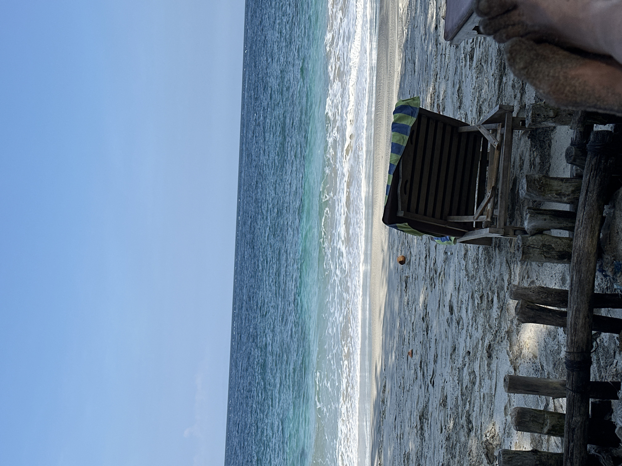 A beach scene in Manggis, Indonesia, features a wooden lounge chair with a blue and green striped towel draped over it, positioned on a sandy beach. The ocean, with clear blue and turquoise water, stretches out to the horizon under a bright, clear sky. Small waves gently lap the shore. In the foreground, a rustic wooden fence made of branches and logs separates the beach area. Two coconuts are visible on the sand near the lounge chair. The lower right corner of the image shows a person's feet covered in sand, suggesting they are relaxed on the beach. The lighting indicates it is daytime, with shadows cast on the sand.