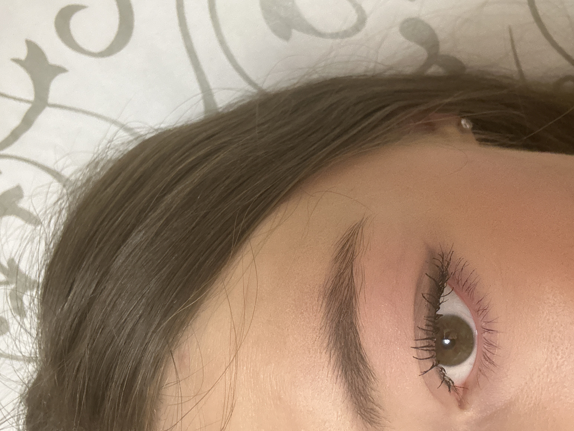 The image shows a close-up view of a person's face, specifically the right side. The person has brown hair, an eyebrow with defined strokes, and an eye with brown iris and long eyelashes enhanced with mascara. The skin appears to be light-toned. A small piercing is visible on the upper ear. The background is a patterned fabric, possibly a bedsheet. The image does not contain location-specific information related to Kazan, Russia.