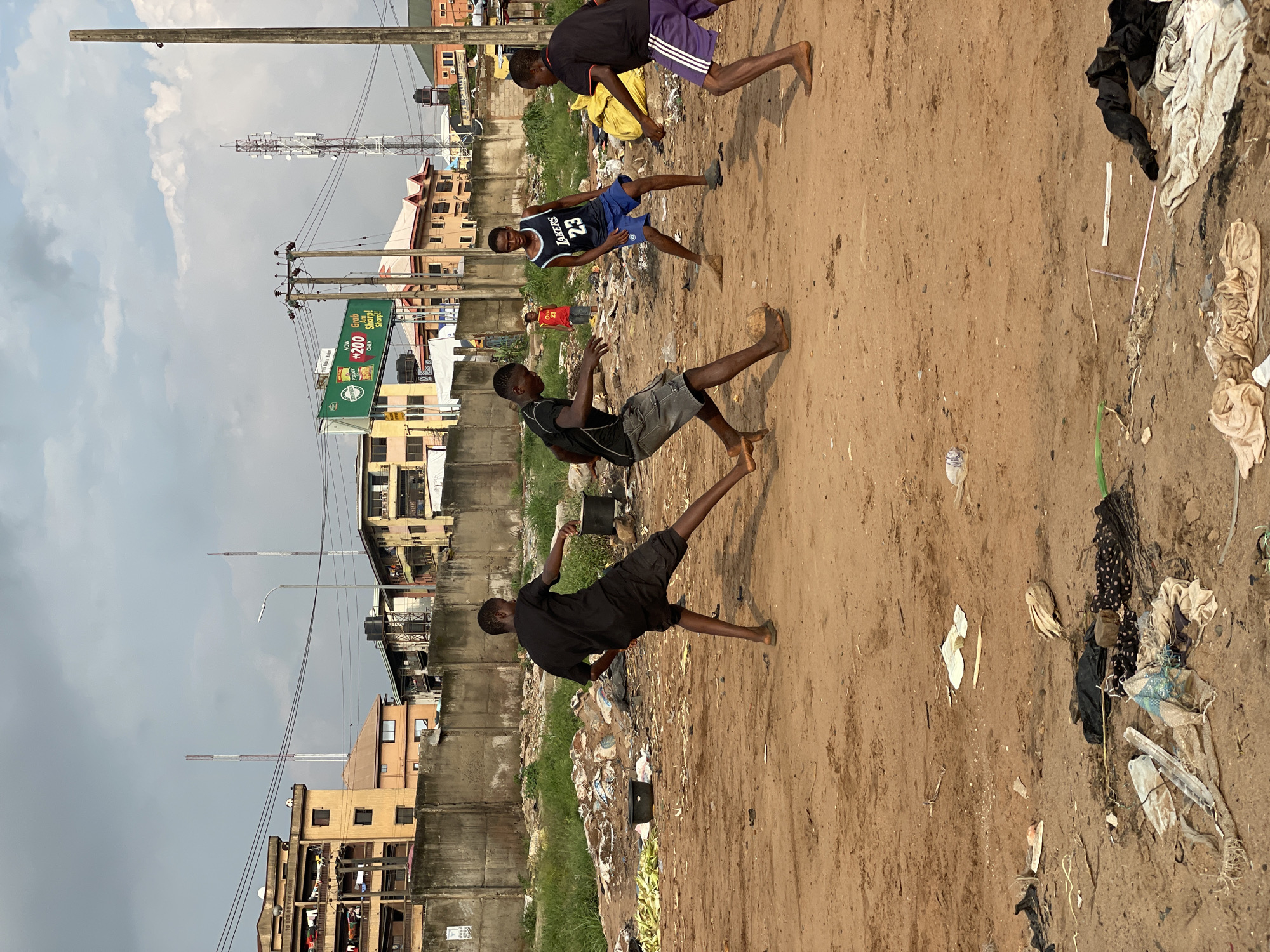 This image depicts an outdoor urban scene in an city within Nigeria, under an overcast sky. In the foreground, four young males are playing football on a reddish-brown dirt field extensively covered with various litter, including plastic, fabric, and organic waste. One player, wearing a black t-shirt and dark shorts, is positioned on the left, engaging with a light brown, makeshift spherical ball with his right foot. To his right, another player in a dark blue "LAKERS 23" jersey and blue shorts stands observing the ball. A third player, in a black t-shirt and purple striped shorts, approaches from the right. A fourth player in black and grey shorts is partially visible near the ball. In the mid-ground, a grey concrete wall stretches horizontally, behind which a younger male in a red t-shirt is visible. Beyond the wall, a dense urban landscape features multiple multi-story buildings of varying colors and conditions. The background also includes numerous utility poles with power lines, a streetlamp, several communication towers, and a green billboard advertising "₦200 ONLY" and "Grab Am Sharp!". The sky is uniformly cloudy.