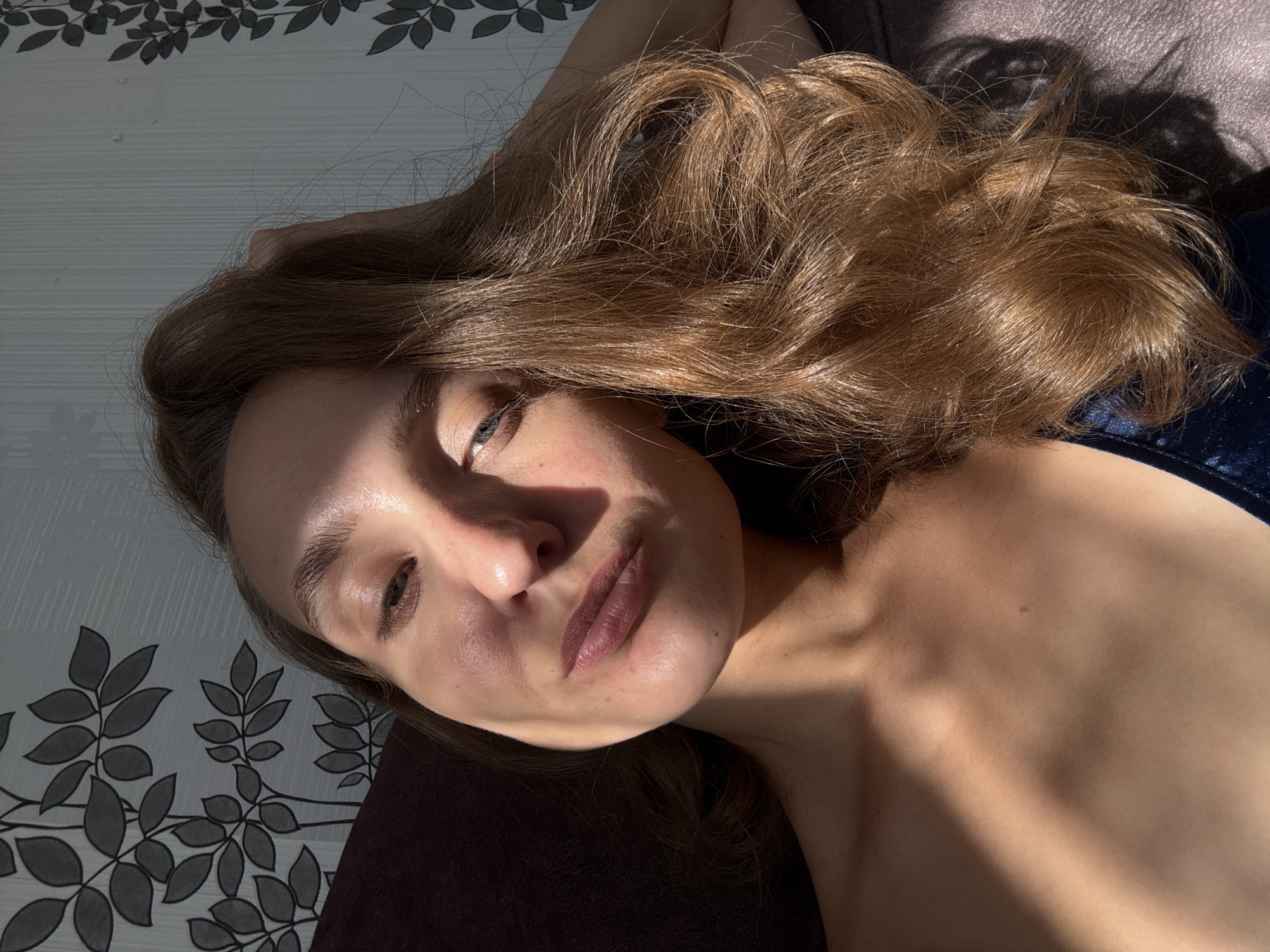 The image is a selfie of a young woman with light brown hair. She is lying down, with her head resting on a dark-colored surface, possibly a couch or bed. Sunlight streams across her face, creating strong shadows and highlighting her features. She appears to be indoors, as indicated by the patterned wallpaper visible in the background. The wallpaper features a dark floral or leaf design against a light gray textured background. The woman is wearing a dark blue garment, possibly a dress or top, with a subtle sheen. Her gaze is directed towards the camera, and her expression is neutral or slightly contemplative. No specific location in Sharkan, Russia, can be identified from the visual information provided in the image.