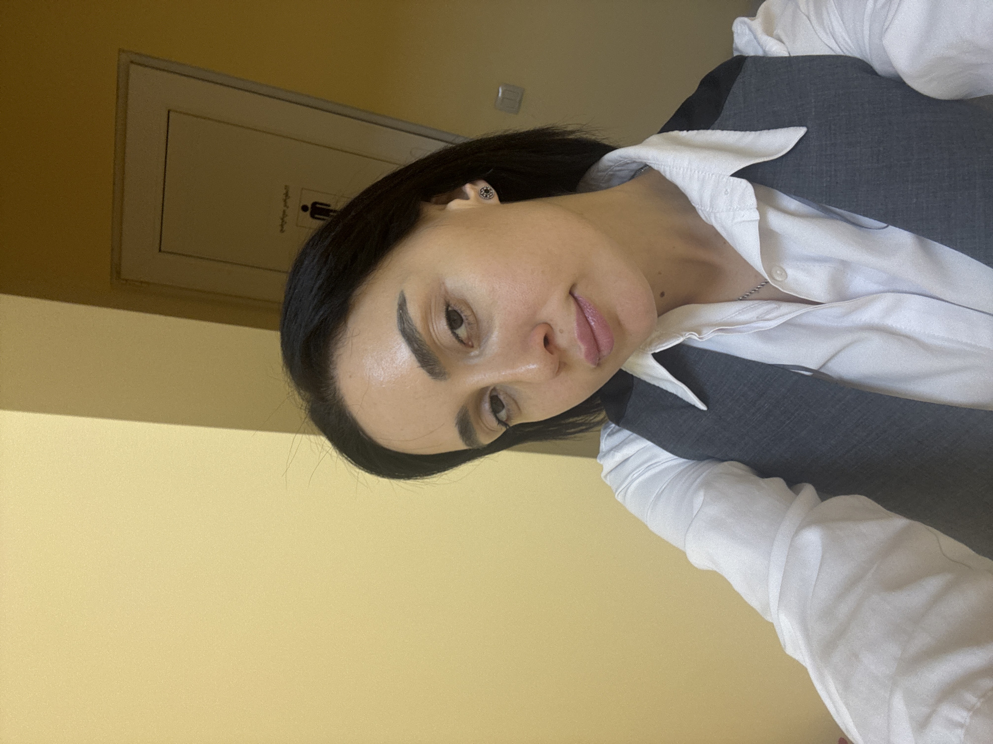 A woman is photographed indoors with a light yellow wall as the background. The room appears to be a public space, as a door with a restroom symbol is visible on the wall. The woman has dark hair and is wearing a white collared shirt with a grey vest over it. She is positioned off-center in the frame, taking up the majority of the shot. The photo is taken from a slightly elevated angle, with the camera tilted. The precise location in Tbilisi, Georgia, is not discernible from the visual information alone.