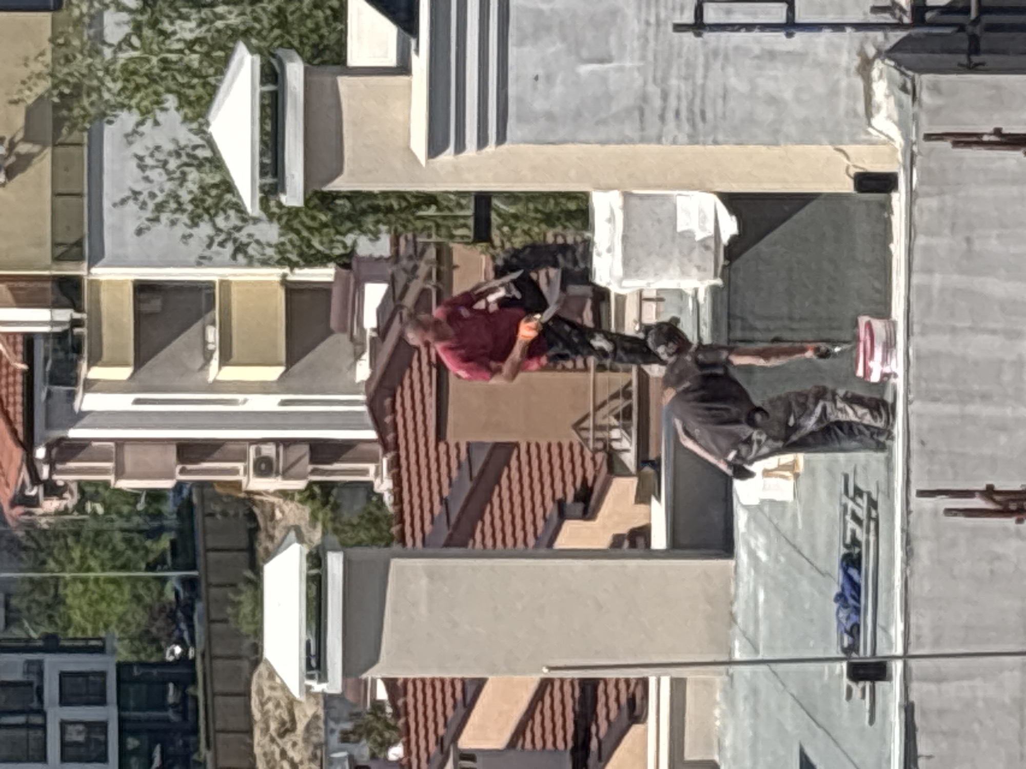 An elevated, slightly tilted angle captures two individuals on a rooftop in Plovdiv, Bulgaria. One person, wearing a red shirt and dark pants, holds an orange object. The other individual, dressed in a dark shirt and camo pants, paints the roof with a roller. A bucket is nearby. The surrounding environment shows a dense urban setting with residential buildings of various heights and roof types, including both flat and pitched roofs with terracotta tiles. A tree with dense foliage is visible amidst the buildings.