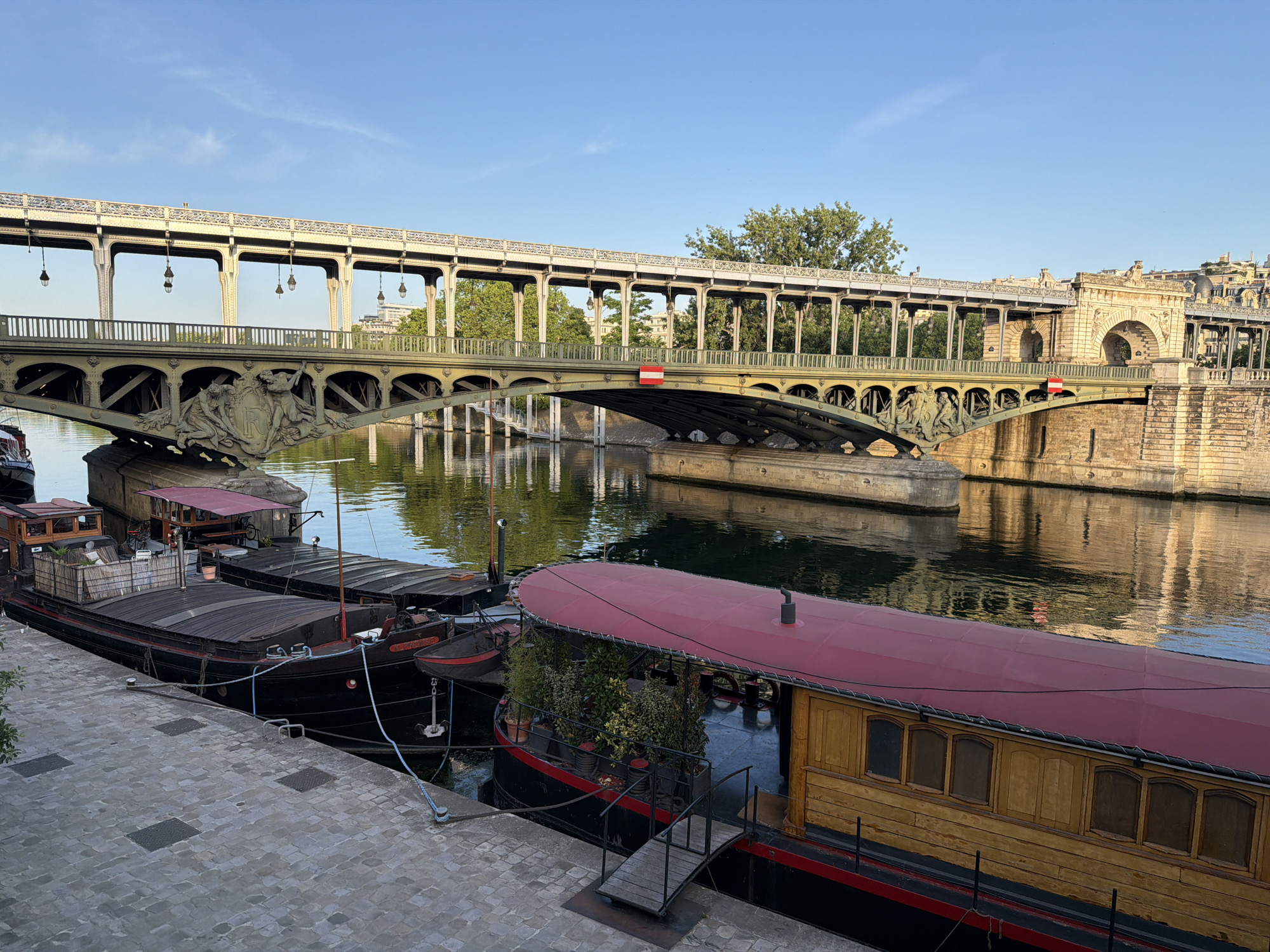 The image captures a Parisian scene with a focus on the Pont de Bir-Hakeim, a notable bridge in Paris, France, crossing the Seine. The bridge is characterized by its two levels: a lower level for vehicles and pedestrians, and an upper level which carries Line 6 of the Paris Métro. The structure has distinct architectural elements, including decorative sculptures and lampposts.
Below the bridge, on the river Seine, a series of barges or houseboats are docked along the riverbank. These boats are of various designs, some with living spaces visible, including wooden structures with windows and a barge covered with a red-colored roof. Plants, some potted, are visible on at least one of the boats. The reflection of the bridge and surrounding architecture can be seen in the water.
The riverbank is paved with light-colored stone. The sky above is clear, with shades of blue.