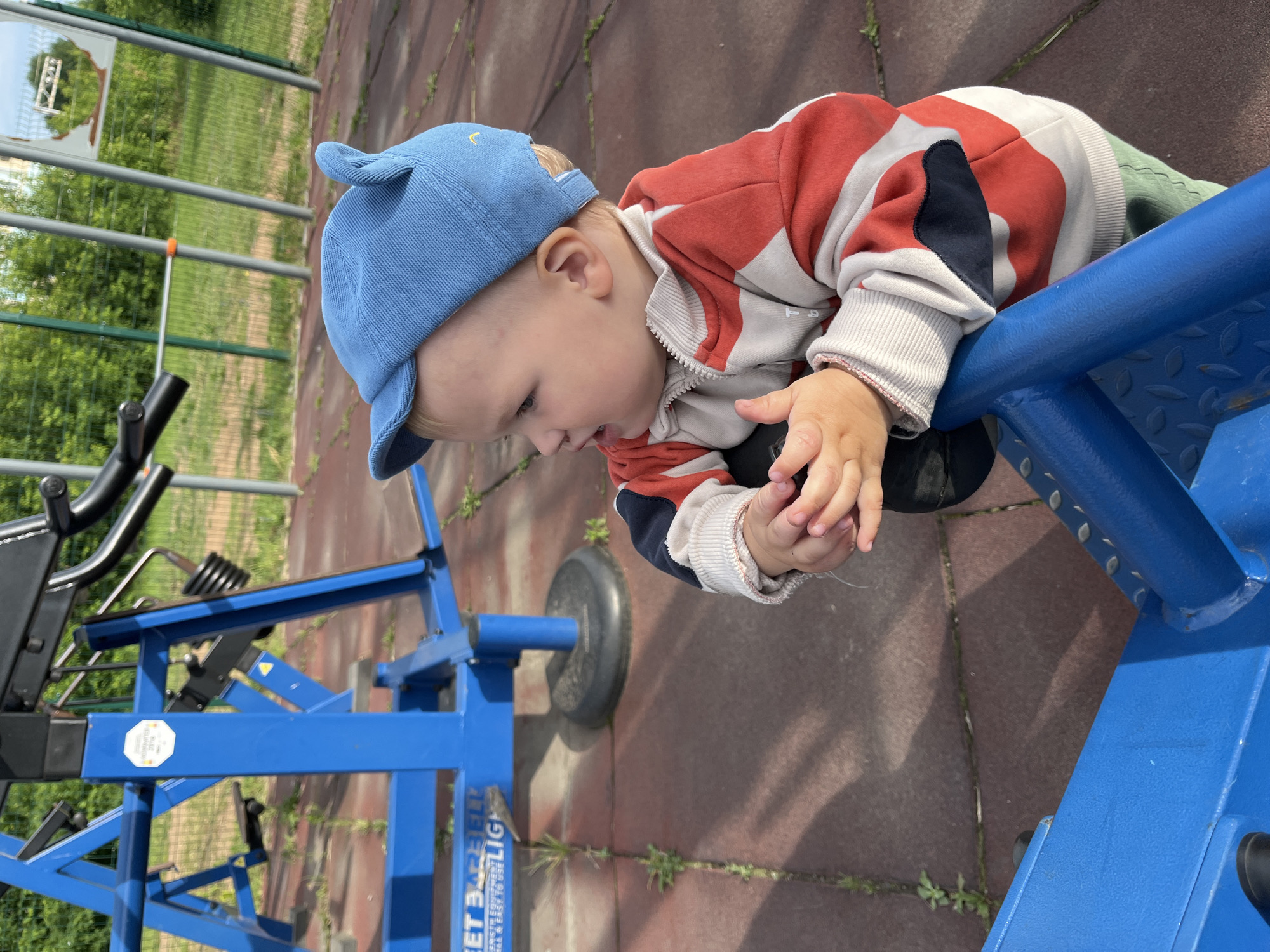 An outdoor children's playground, possibly located in the city of Chaikovsky, Russia, features a young child in a blue cap and a red and white striped jacket. The child is leaning on a blue metal structure, which appears to be part of a piece of playground equipment. The ground is a reddish-brown rubberized surface, with patches of grass growing through. In the background, a green fence encloses the playground, with trees and foliage visible beyond it. The lighting suggests it is daytime, with sunlight casting shadows.