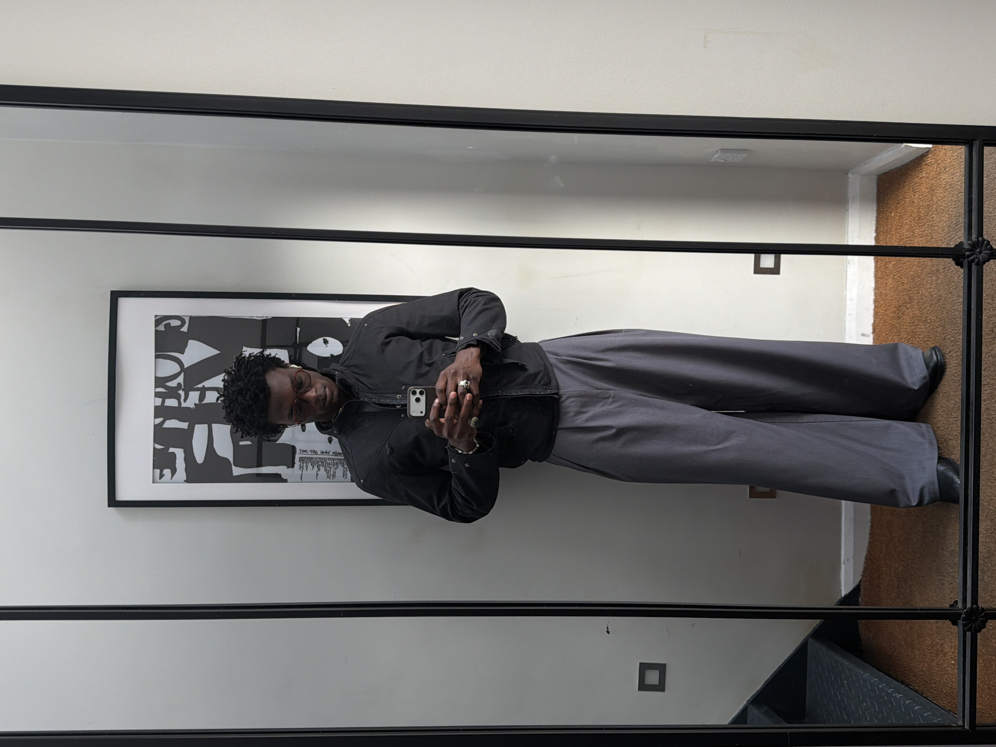 A man is taking a selfie in a large mirror in an indoor setting. The mirror is framed in black metal with decorative elements and reflects the hallway. The man is wearing a dark jacket and gray pants, holding a smartphone. He has short, dark hair and is wearing glasses and rings. Behind him, hanging on a white wall, is a framed piece of artwork. The environment includes neutral-colored walls, a dark frame around the mirror, and glimpses of a stairway and flooring within the mirror's reflection. There is a dark square electrical outlet on the wall below the picture frame. Based on the architectural style and interior elements, the location might be a residential space in London, United Kingdom.