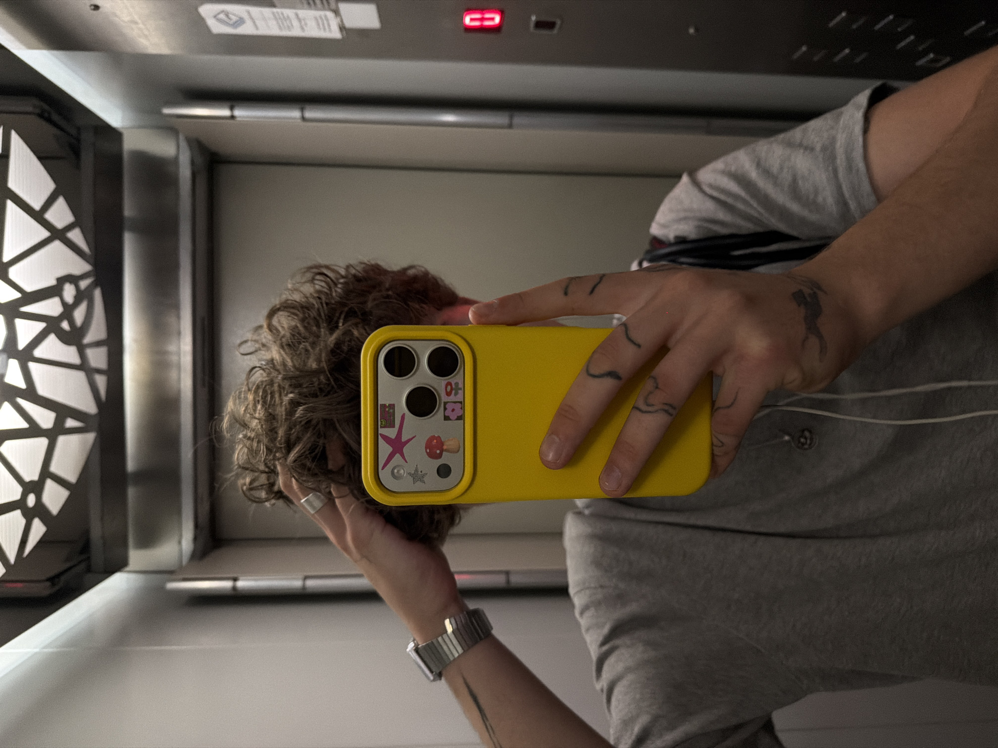 A man takes a selfie in an elevator. The elevator has a modern interior with geometric lighting and a digital display panel. The man is wearing a gray shirt, a watch, and a ring. He holds a yellow phone case adorned with stickers. The exact location within Vracar Urban Municipality, Serbia, cannot be determined from the image alone.