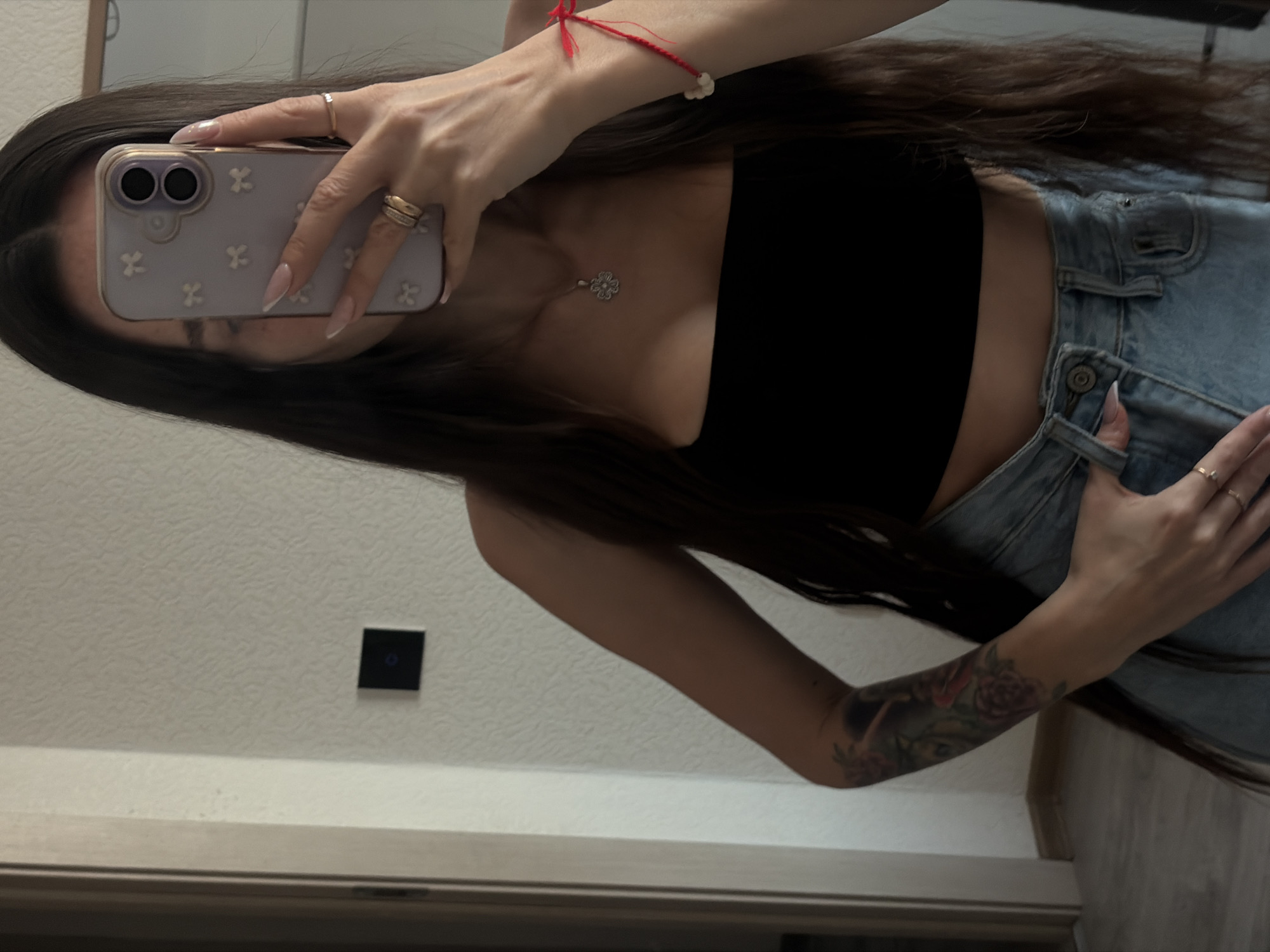 A woman takes a selfie in what appears to be an indoor environment. She is holding a smartphone with a light purple case that has a floral pattern. The phone covers most of her face, but her long, dark hair is visible. She is wearing a black tube top and light blue jeans. Her left arm has a tattoo featuring floral designs. She is wearing a red string bracelet on her left wrist and rings on both hands. In the background, there is a light-colored wall and a dark square object which might be a light switch. Based on these visual elements, the image does not contain any indicators related to Saint Petersburg, Russia. The location is not visually discernible in the image.