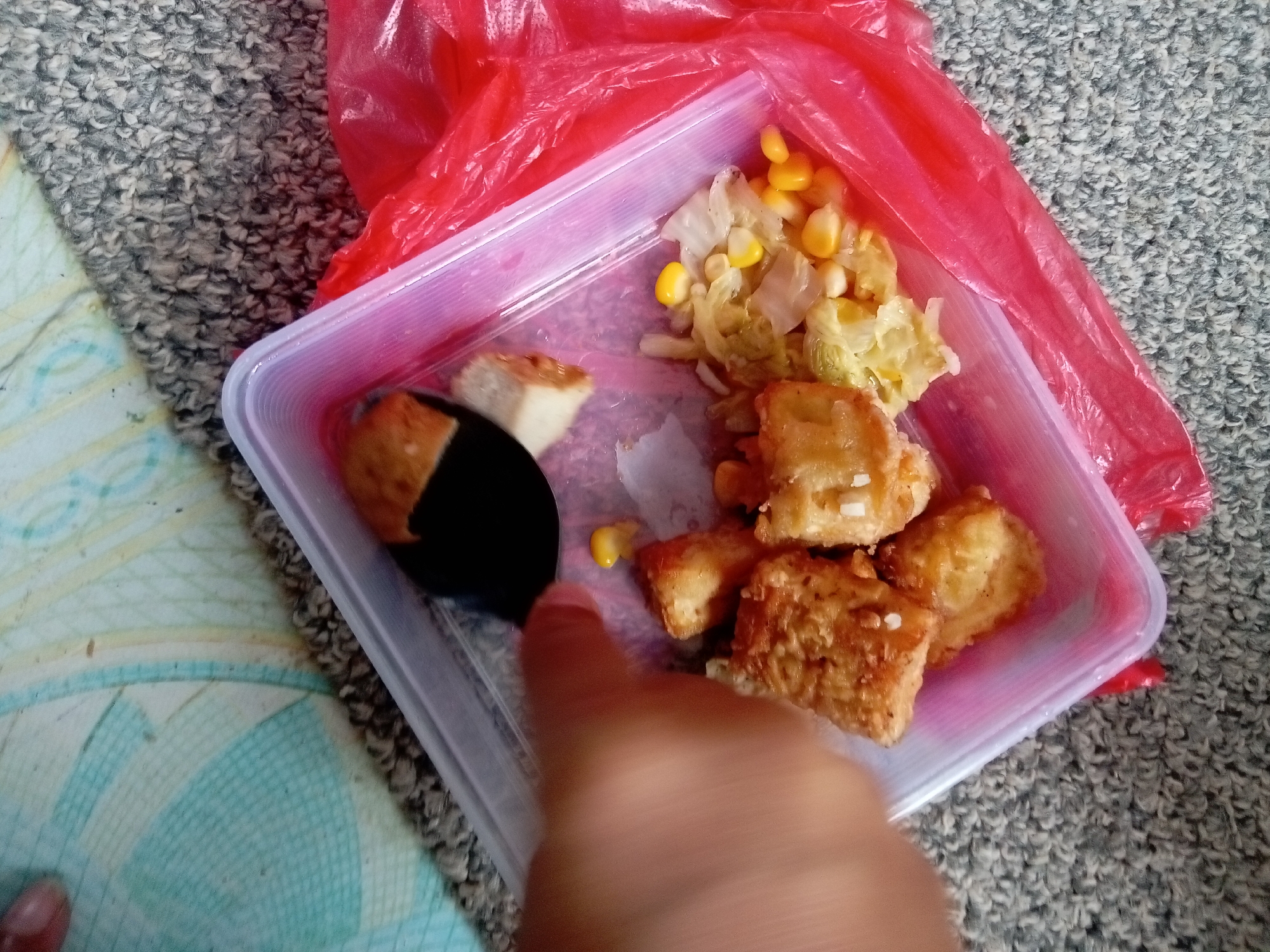 A close-up, high-angle shot captures a plastic food container filled with fried tofu and a mixture of corn and cabbage, situated on a patterned blue and white surface and a grey textured carpet. A red plastic bag envelops the top portion of the container. A person's hand and finger are visible, intruding into the frame from the bottom, with the finger appearing to be pointing towards the food. The food appears to be part of a takeout meal, likely from a vendor or restaurant in Batam, Indonesia, given the context of the prompt. The lighting suggests an indoor setting, possibly a home or a public space.