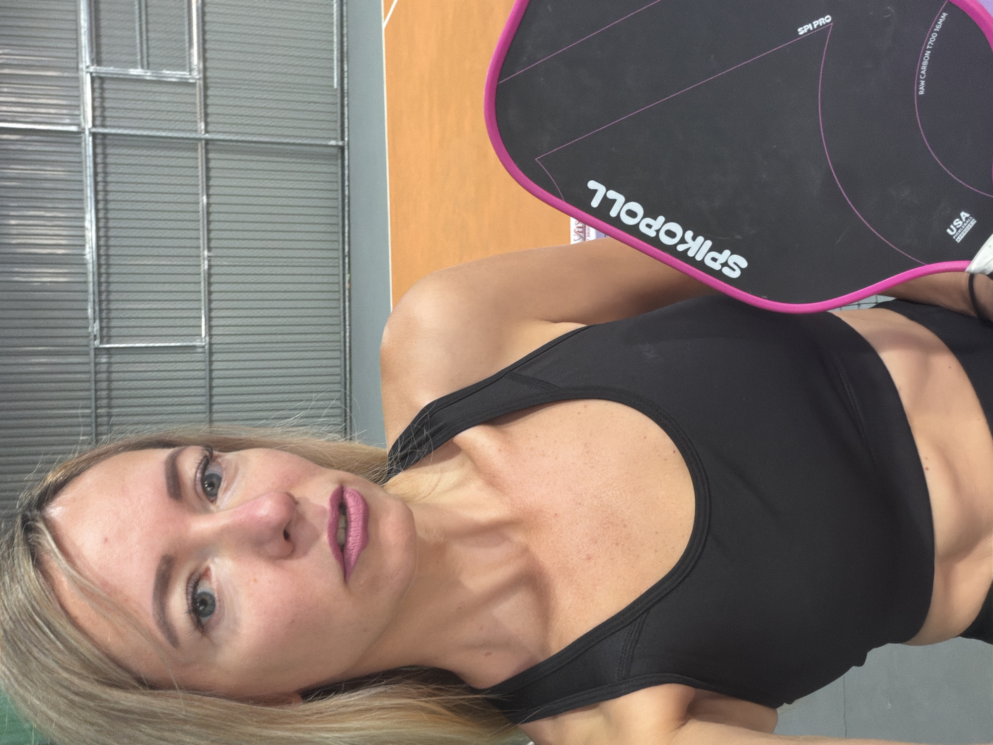 A young Caucasian woman with blonde hair and blue eyes poses for a selfie in an indoor pickleball court. She is wearing a black sports bra and has purple lipstick on. She is holding a black pickleball paddle with pink trim and white lettering that reads "SPIKOPOLL". The background consists of a gray corrugated metal wall and an orange pickleball court with gray flooring. The image appears to have been taken in Da Lat, Vietnam, where the woman is likely participating in a sporting event or recreational activity.