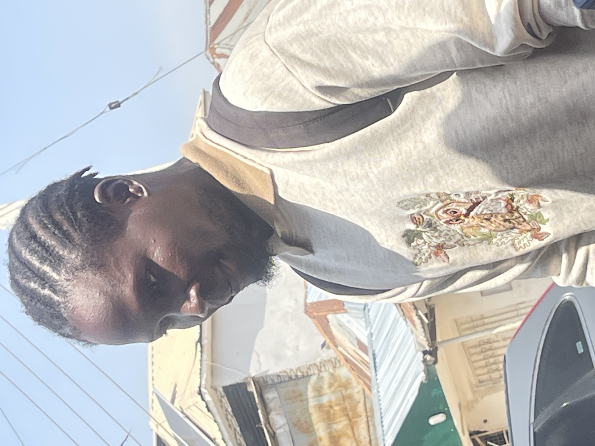 The image shows a medium shot of a Black man with braided hair, facing to his right, looking downwards. He is wearing a light-colored crewneck sweatshirt with a tan collar and a graphic design of an owl with floral elements on the left chest. A dark backpack is visible over his shoulders. The background consists of a bright blue sky with overhead wires, and buildings with corrugated metal roofs and some white structures. The scene appears to be outdoors in a somewhat urban or densely populated residential area. The lighting suggests it is daytime. The overall impression is of a person in an everyday setting, possibly on a street or walkway. The image's composition and focus are on the man, with the background slightly out of focus.