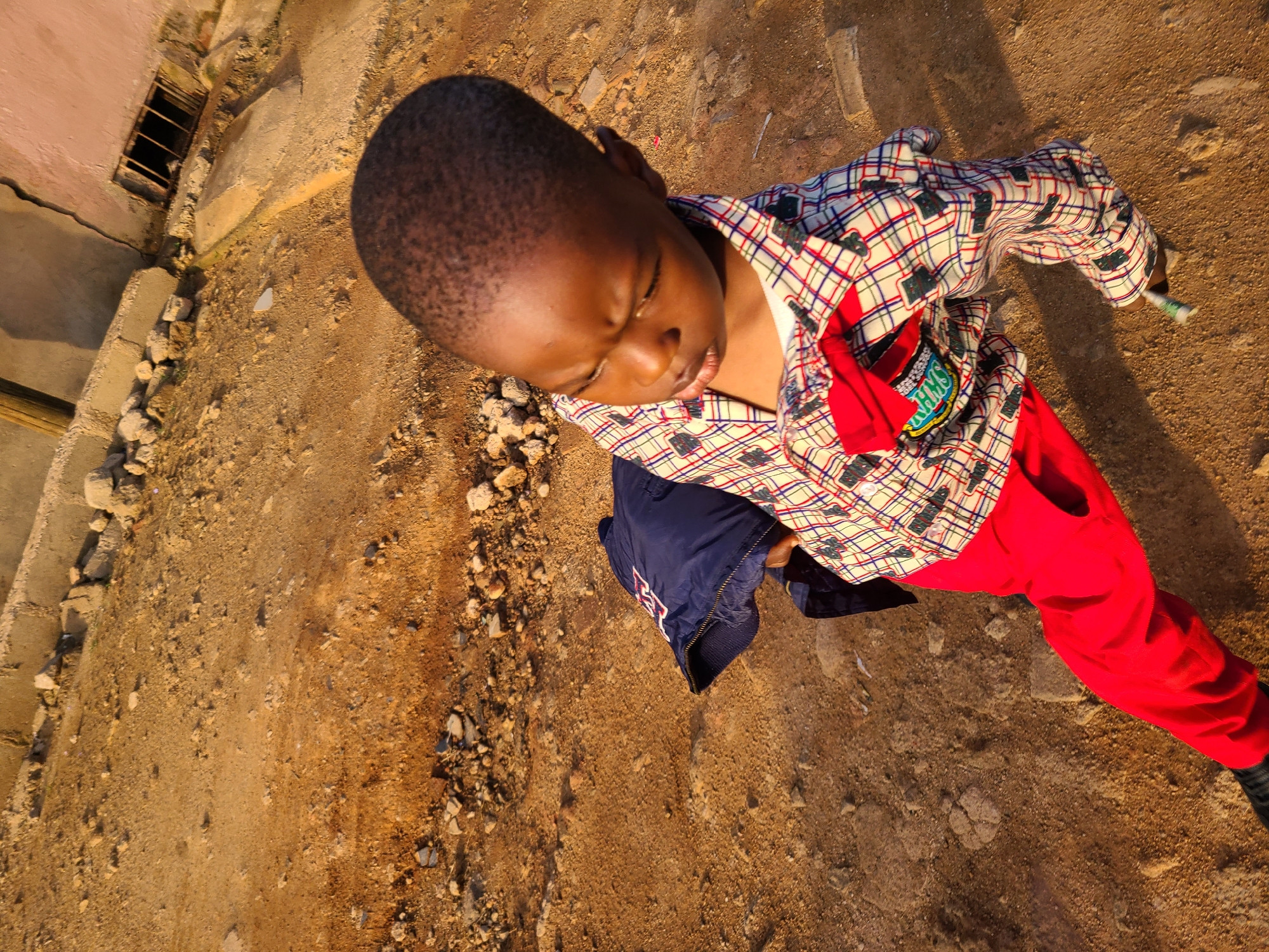 A young boy is pictured outdoors, standing on a patch of dirt and gravel. He is wearing a plaid shirt with a red bowtie, red pants, and a dark blue jacket. The background shows a rough, unpaved surface with scattered rocks and debris. In the upper right corner, a portion of a building with a metal grate and cracked concrete is visible. The lighting suggests it is either early morning or late afternoon, casting long shadows. The environment appears to be an undeveloped area within a city, possibly in Jos, Nigeria, given the context. The boy's posture and gaze suggest he is looking down and slightly to his left.