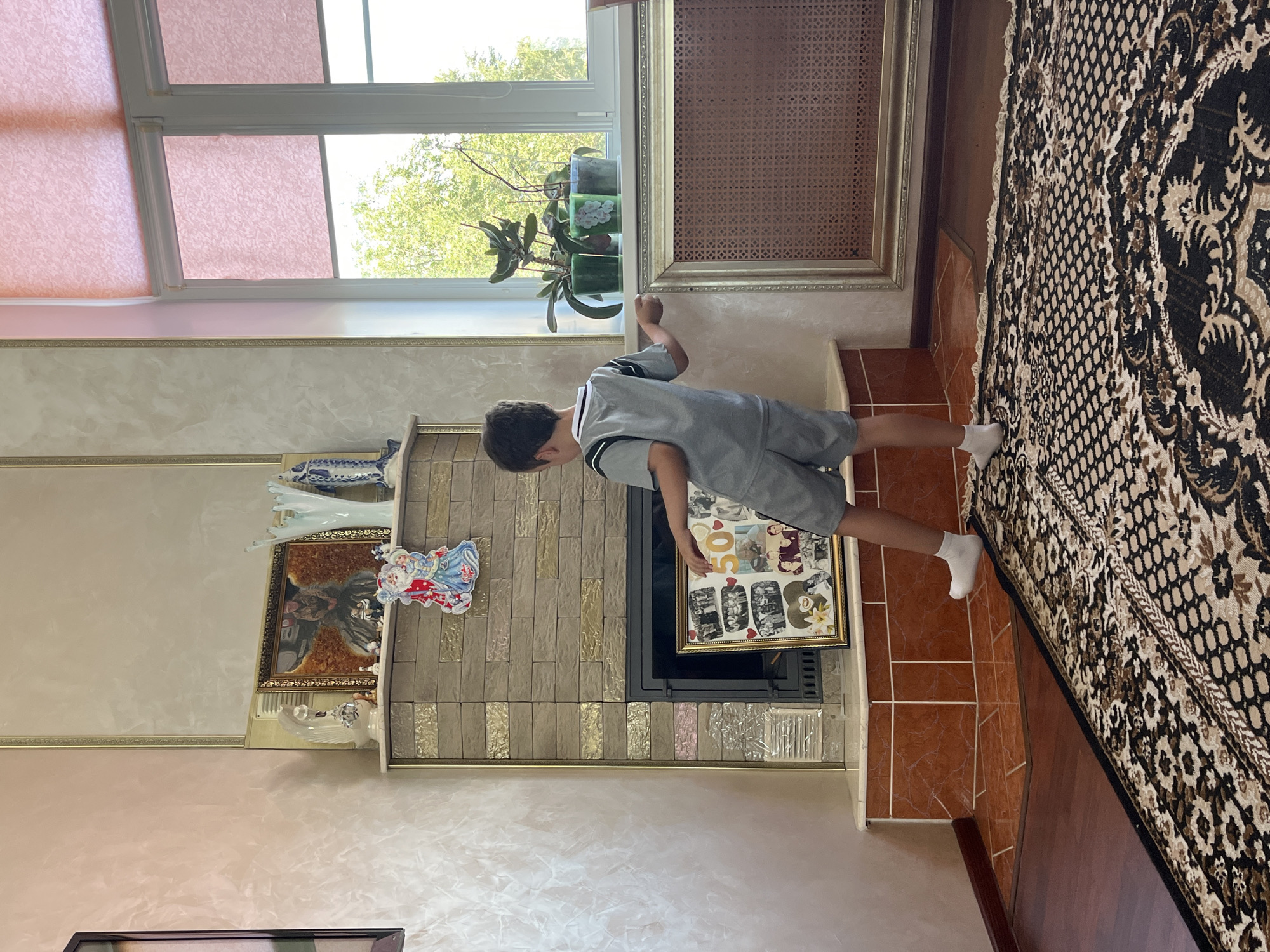 A young boy in a grey t-shirt and shorts stands in a room, facing a fireplace. He is holding a framed collage of photographs in his right hand. The collage appears to be a collection of family photos, with a prominent "50" in the center. The fireplace is made of stone and has a dark metal insert. Above the fireplace, there are several decorative items, including figurines and vases, and a framed painting. To the right of the fireplace is a large window with pink roller blinds, through which trees and greenery are visible. In front of the window, on the windowsill, are several potted plants. A patterned rug covers the wooden floor in the foreground. The room has a light-colored wall and a radiator with a decorative gold frame. The lighting suggests it is daytime. The boy is wearing white socks.