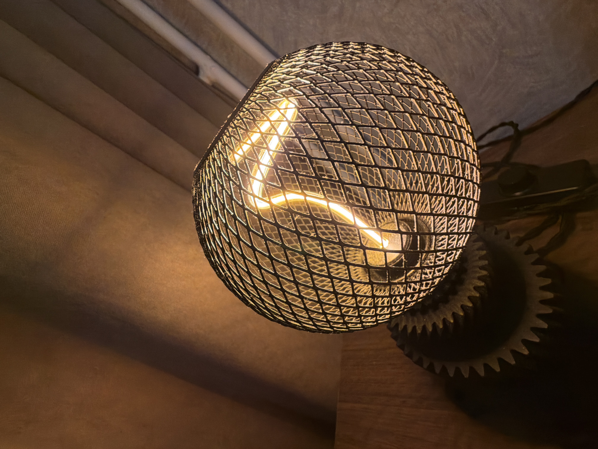 The image displays an illuminated light fixture, focusing on a bright light bulb housed within a black, diamond-patterned metal mesh shade. The bulb emits a warm, vibrant yellow light, highlighting its distinct curved filament, which forms a shape resembling an 'L' or a hook. The mesh shade casts intricate diamond-shaped patterns of light and shadow onto the surrounding surfaces.
To the upper left, the background consists of a soft, textured surface with prominent horizontal folds or stripes, suggesting closed curtains or blinds in muted brownish-grey tones, softly illuminated by the lamp. The lower right portion of the image reveals a dark wooden surface, reflecting some of the warm light. On this wooden surface, a dark, gear-shaped object with clearly visible teeth is partially silhouetted, casting its own shadow. A dark electrical cable or wire is also visible trailing behind the light fixture. The overall scene suggests an indoor setting, likely in the evening or a dimly lit room, with the artificial light from the lamp serving as the primary light source. No discernible text is visible in the image.