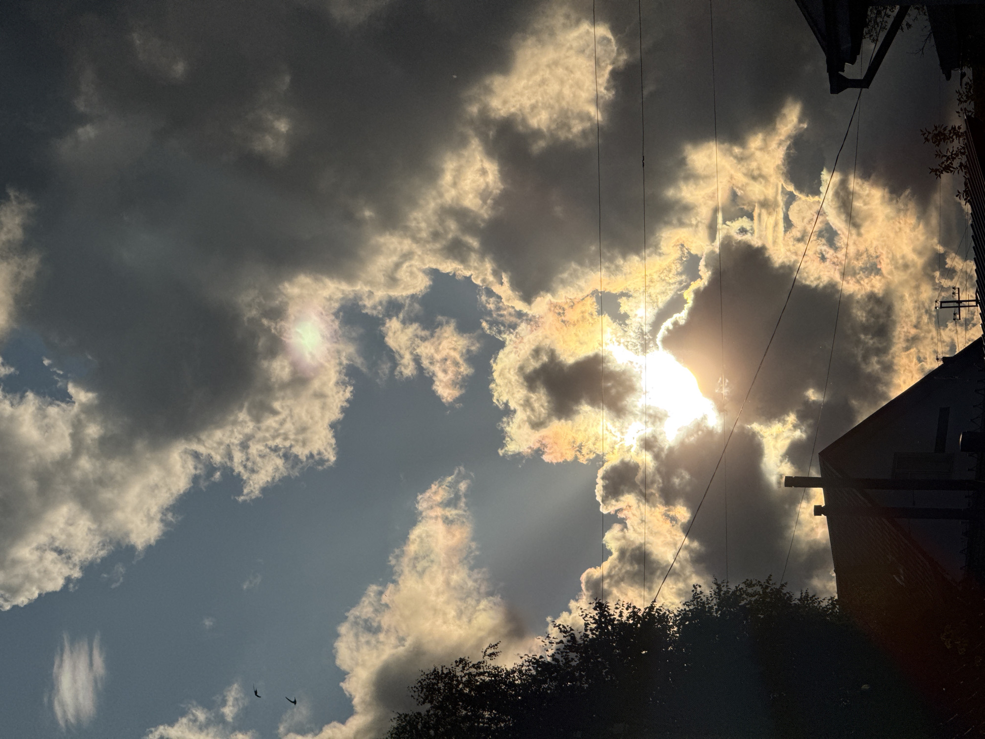 The image depicts a dramatic sky over Ветлужский, Russia, with a storm brewing. Dark, heavy clouds dominate the upper portion of the frame, contrasted by patches of bright, illuminated clouds that suggest the sun is breaking through. Streaks of sunlight pierce through the clouds, creating a theatrical effect. In the foreground, the silhouette of a house with a prominent chimney is visible, along with power lines stretching across the sky. The overall atmosphere is one of impending weather change, with a blend of darkness and light. Two birds are seen flying in the distance.