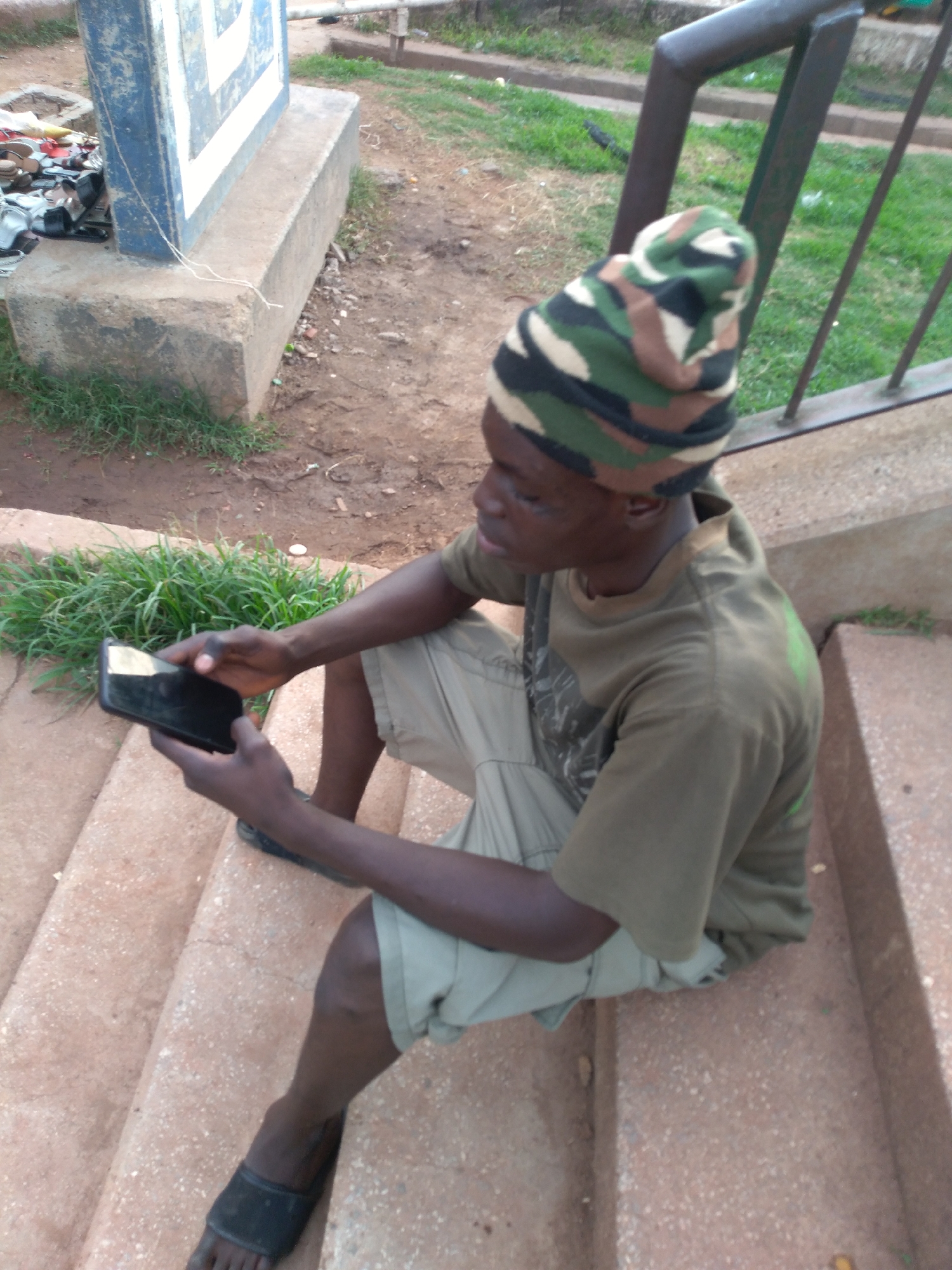 A young African male, wearing a camouflage beanie and a green t-shirt with light-colored shorts, sits on concrete steps outdoors. He is holding a black smartphone in his hands, with his left index finger appearing to touch the screen. The environment appears to be a public space, with worn concrete steps and patches of grass visible. In the background, a metal railing and the base of a concrete structure with a blue and white design can be seen. There are also some discarded items, including footwear, near the concrete structure. The scene is not clearly identifiable as Gura Top, Nigeria, but the overall setting suggests an urban or semi-urban outdoor location.