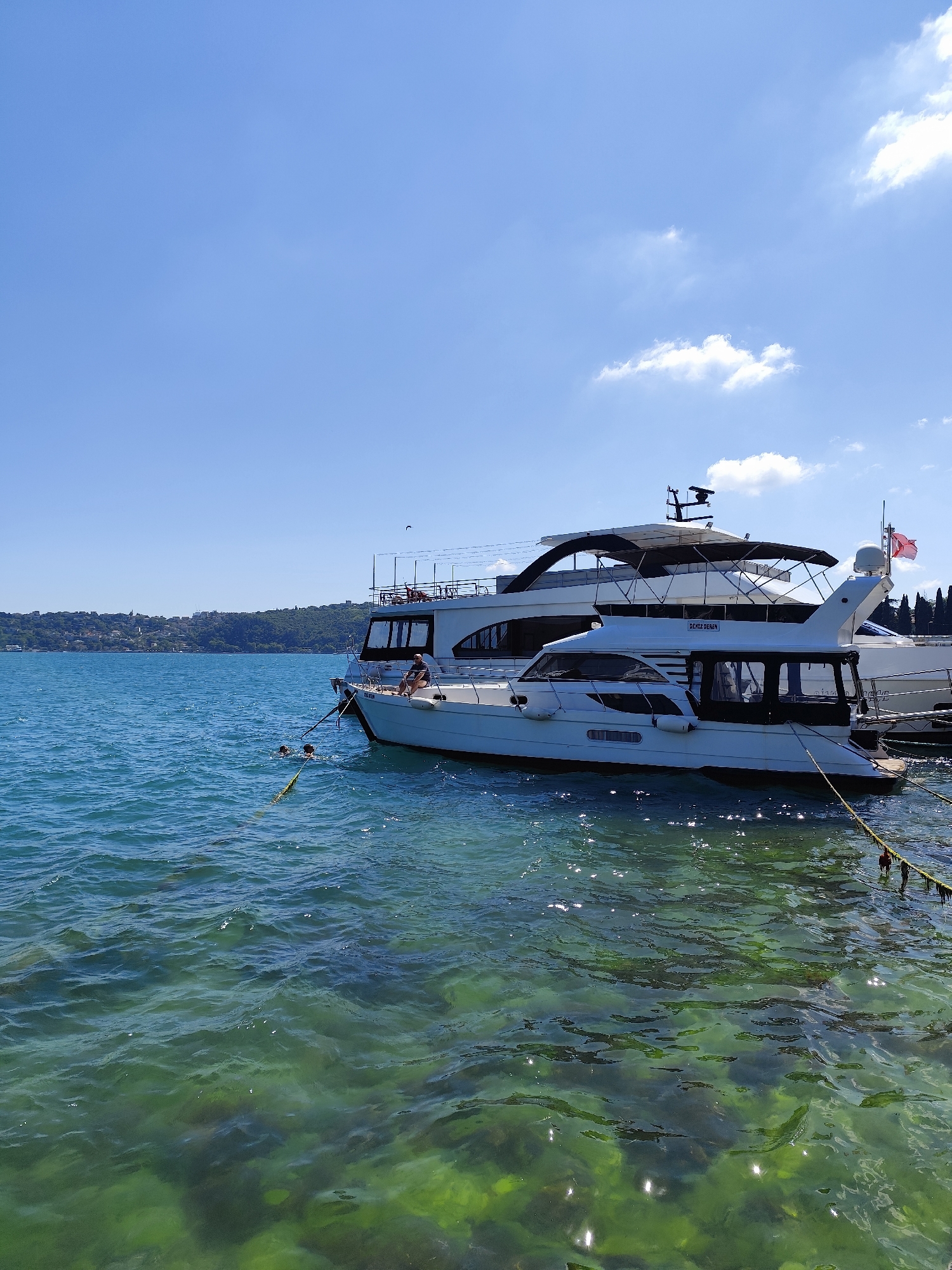 Two white yachts are moored in clear, green water under a bright blue sky with scattered clouds. The yacht in the foreground is smaller and has a black canopy. A man is seated on the bow of this yacht. In the water next to the yacht, two individuals are swimming and holding onto a rope attached to the boat. The larger yacht behind it is mostly obscured. The background shows a tree-lined shore with buildings and hills. The scene appears to be a sunny day at a waterfront location.