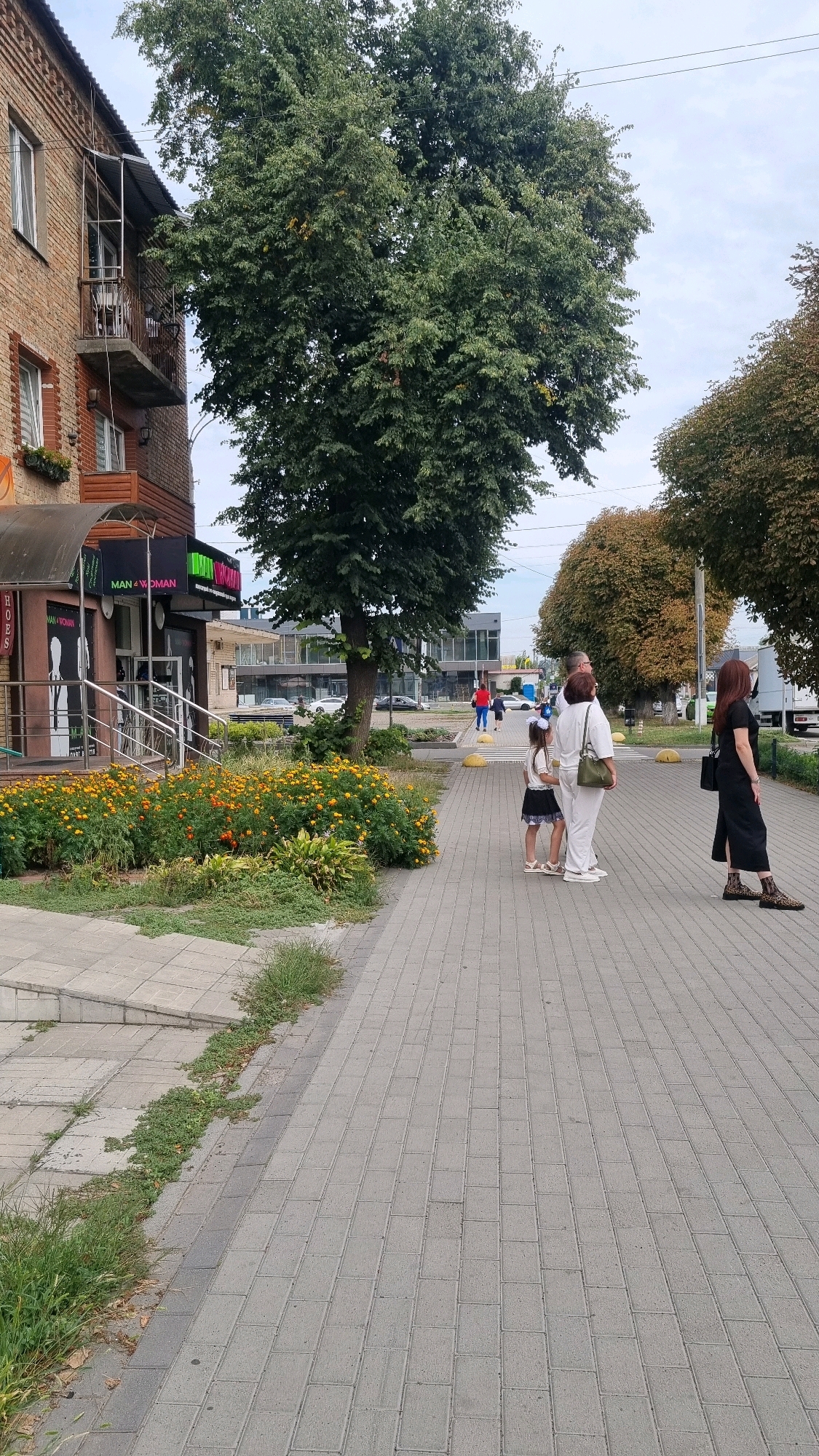 In Smila, Ukraine, on a paved sidewalk next to a brick building with storefronts, a group of individuals are observed. The building features a balcony with metal railings and a canopy over an entrance. In front of the building, a bed of orange marigolds is visible. Further down the sidewalk, two women and a young girl stand together. The older woman is dressed in white, the girl in a black and white dress, and the other woman in a black dress. A fourth woman, also in a black dress, stands further down the sidewalk, facing away from the camera. In the background, trees with green and autumn foliage line the street, along with parked cars and other buildings. The sky is overcast.
