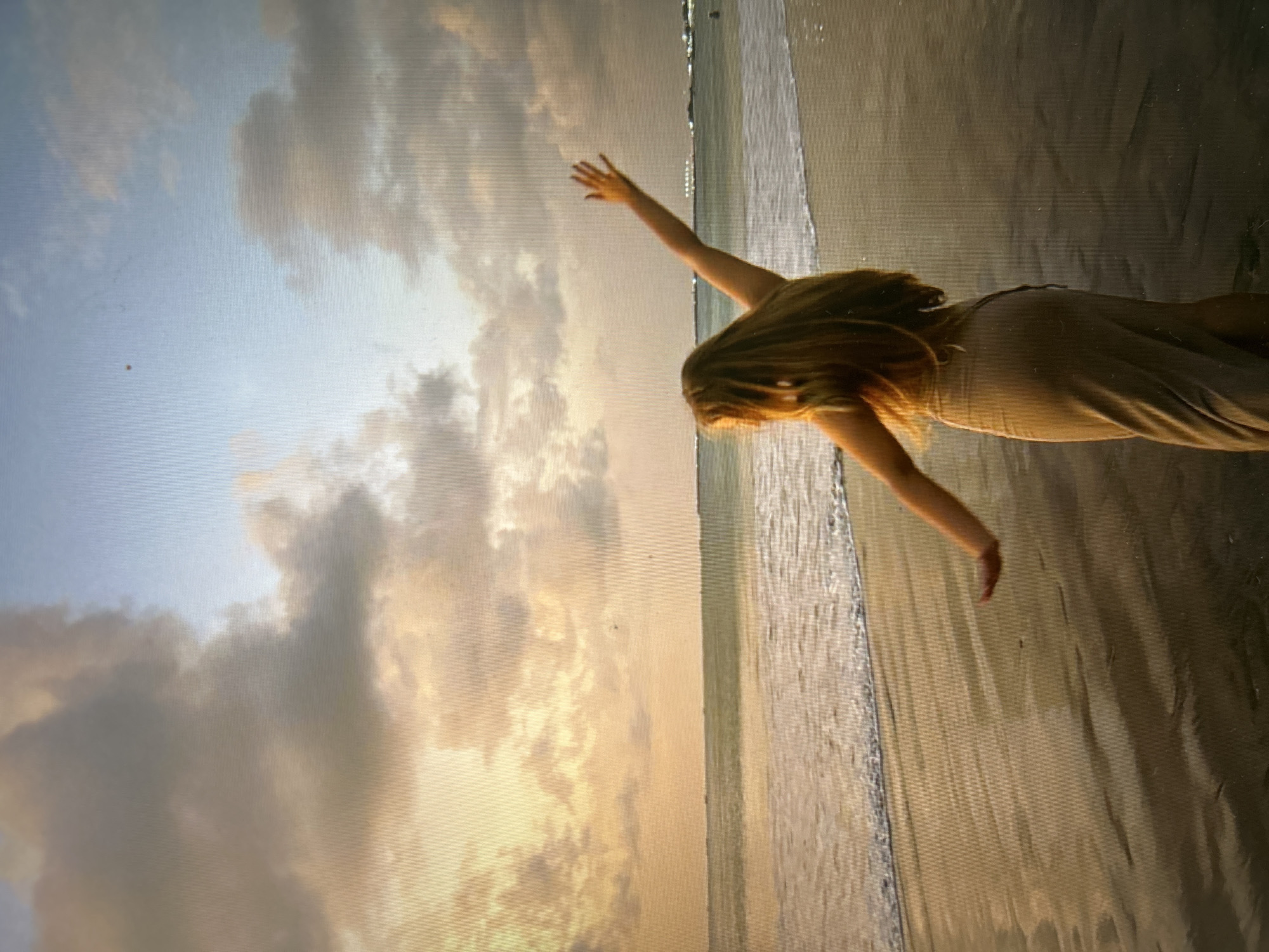 A woman stands on a beach, extending her right arm towards the sky, with her left arm slightly outstretched. She is wearing a light-colored, ankle-length dress. Her long hair cascades down her back. The beach has a sandy texture with the ocean washing gently against the shoreline. The sky is filled with clouds tinged with golden hues of sunset or sunrise. This photo was taken in Kuta Selatan, Indonesia.