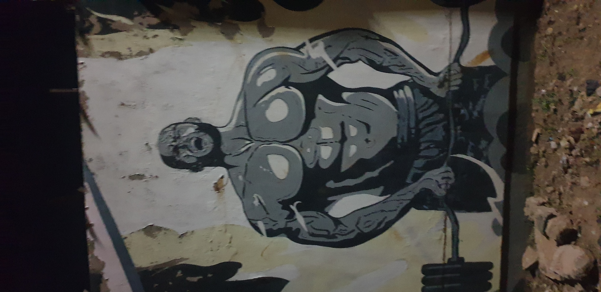 This image, captured in Narayi, Nigeria, displays a mural painted on a distressed light-colored wall with visible peeling paint and discoloration. The mural, rendered in grayscale with white highlights and black outlines, depicts a highly muscular, bald, male figure from the waist up. The figure's head is tilted back, with its mouth open, conveying an expression of intense exertion. It is shown holding a black barbell with weighted plates at waist level, suggesting a lifting action. The figure wears dark shorts. The ground in the immediate foreground is bare dirt, sparsely covered with rocks and small debris. The upper portion of the scene above the wall is a dark, unlit background, indicative of nighttime conditions.