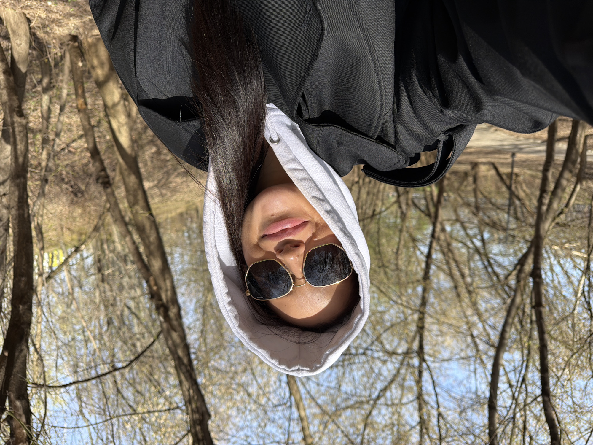 A person with long, dark hair, likely female, is captured in a close-up outdoor portrait against a backdrop of trees. The individual is wearing a white hooded sweatshirt with the hood pulled up, and a black jacket or coat layered over it. Large, gold-framed sunglasses, possibly hexagonal or square in shape, obscure their eyes. The word "Polaroid" is faintly visible on the left lens of the sunglasses. Their lips are slightly parted in a relaxed manner, revealing a subtle expression.
The setting appears to be a wooded area, forest, or park, characterized by numerous bare trees with light brown trunks and branches. This suggests an early spring or late autumn season in a temperate climate. Patches of clear blue sky are visible through the network of branches, indicating fair weather. The scene is brightly lit by strong sunlight, suggesting it is mid-day. The person is looking slightly upwards towards the camera, suggesting a selfie or a low-angle shot. The overall atmosphere is casual and serene, reflecting a moment spent outdoors in a natural environment. The location is Khimki, Russia.