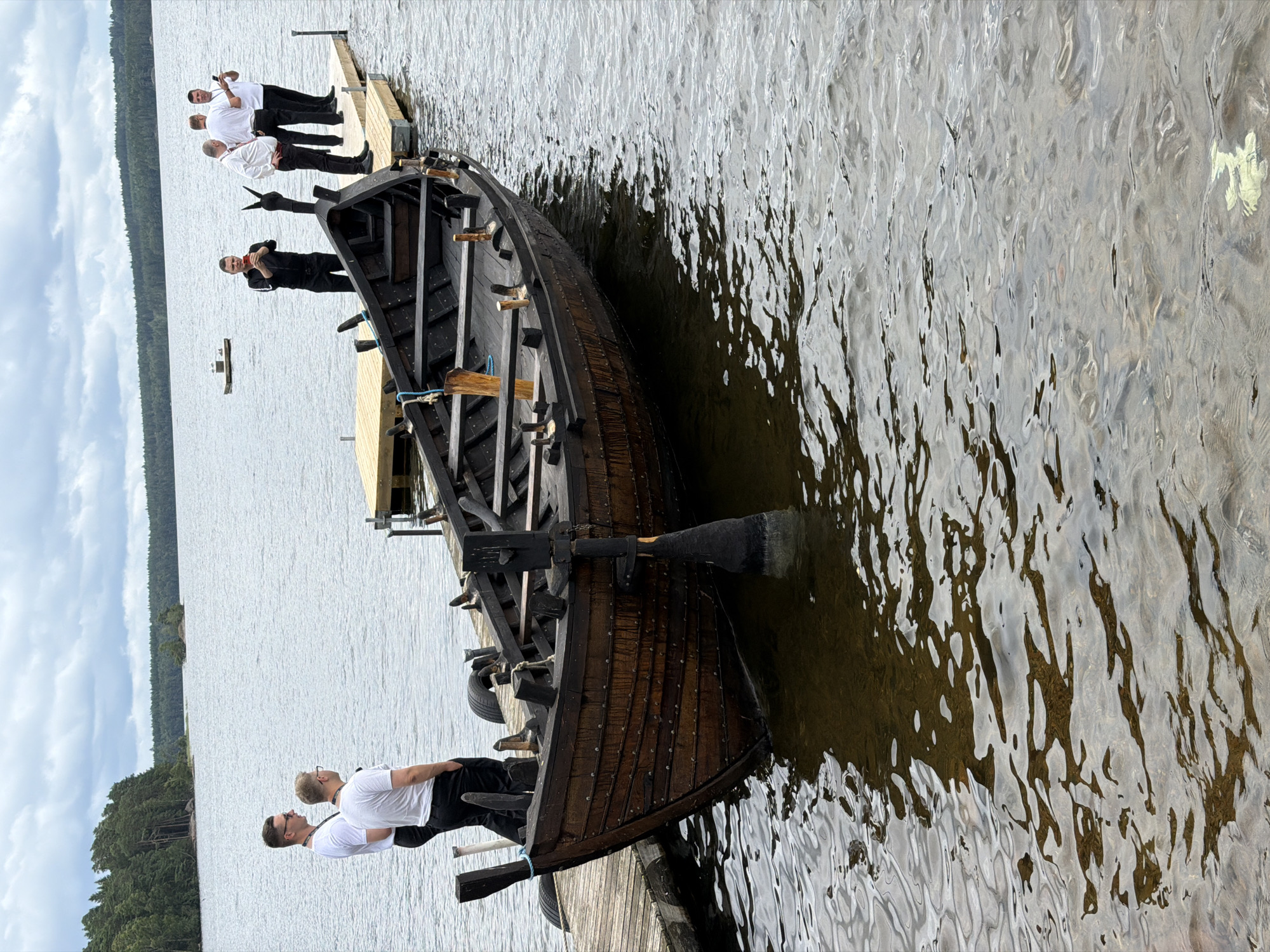 A wooden rowboat is docked at a wooden pier on a lake in Aneby, Sweden. The boat is dark brown and appears to be old, with rivets visible along its hull. An oar is partially submerged in the water next to the boat. Several men are standing on the pier, some are holding phones, and appear to be looking out at the water. The sky is overcast, and the surrounding area is forested.