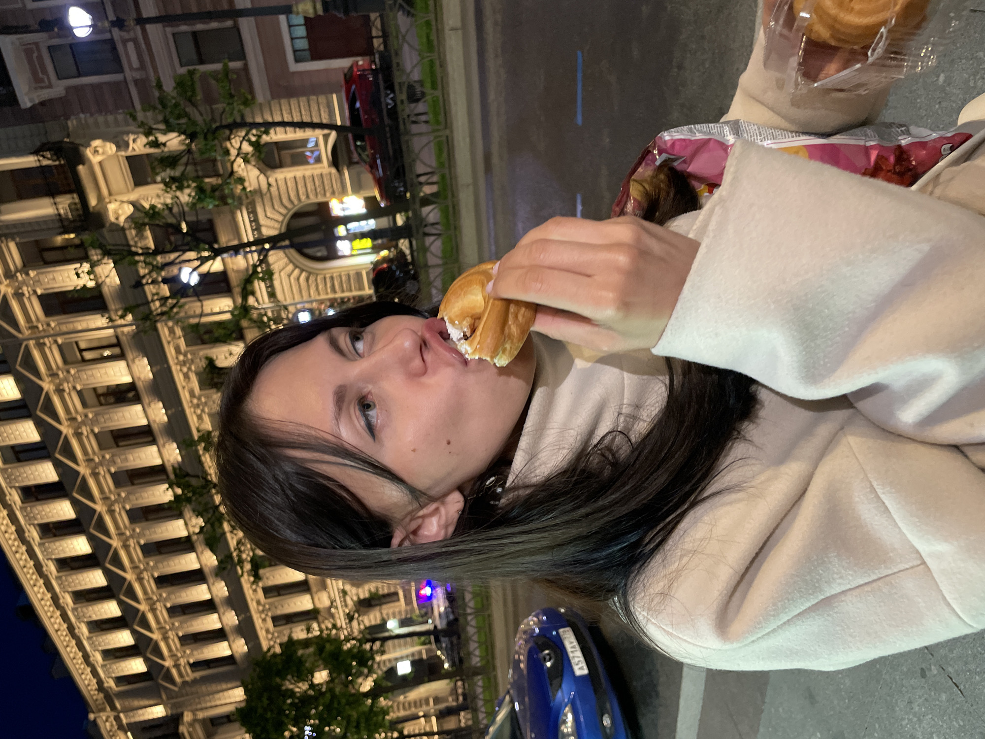 A medium shot shows a young woman eating a pastry on a street in Saint Petersburg, Russia. She is wearing a light-colored coat and has dark hair. The woman is looking to her right and is in the process of taking a bite of the pastry. The background shows a building with illuminated windows, trees, and cars. One car in the foreground is blue and has a license plate that reads "A571AV178". The lighting suggests it is evening or night.