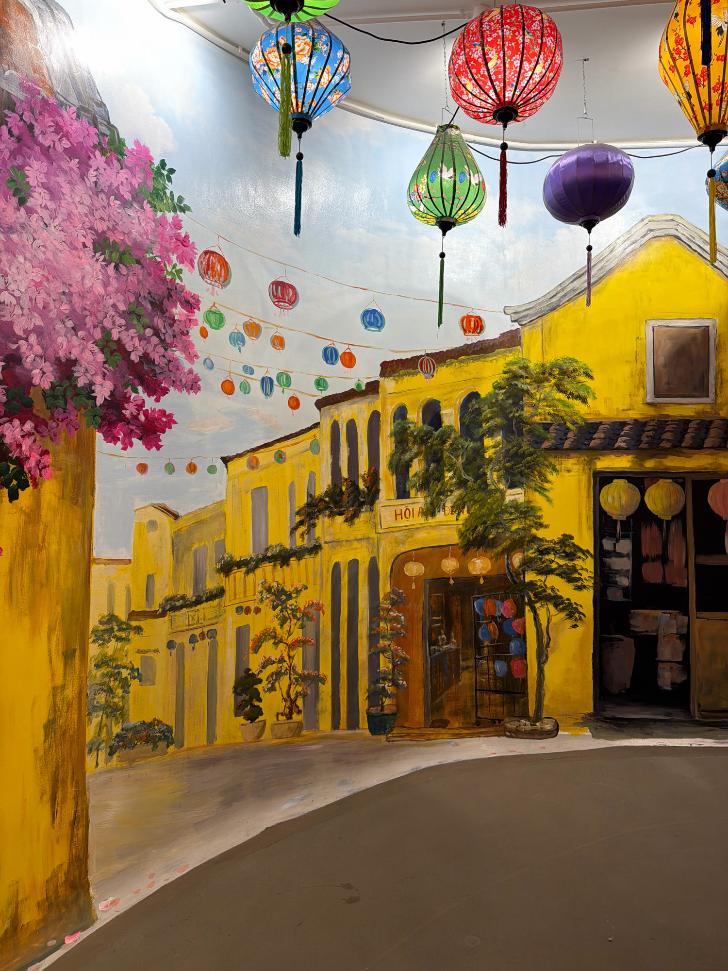 The image depicts a mural painted on a wall, creating the illusion of a street scene in Phú Quốc, Vietnam. The mural features brightly colored buildings, primarily yellow, with architectural elements like arched doorways and windows. Hanging above the street are numerous traditional Vietnamese lanterns in various colors, including red, blue, green, and purple, some adorned with intricate patterns. Lush pink flowers cascade down the left side of the mural, adding a vibrant natural element. In the foreground, a curved road is painted, and a tree with green foliage grows near the buildings. The overall impression is of a lively and festive atmosphere, characteristic of Vietnamese street markets or festivals. There are no people or animals visible in the mural.