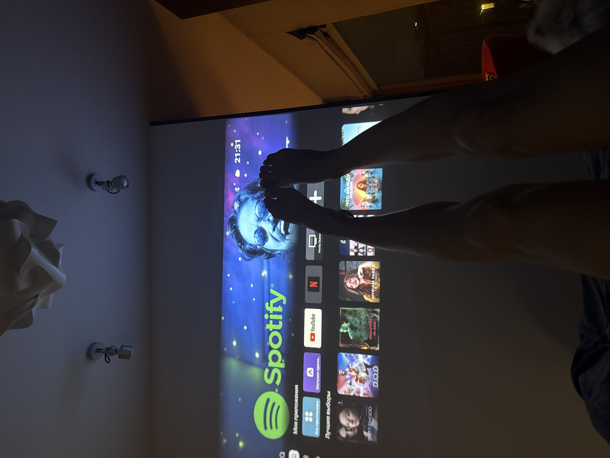 Here's a detailed description of the image:
**Scene:** The image shows a dark interior room, likely a bedroom. A projector displays a screen on the wall. The screen is showing the home screen of a smart TV or media device, with app icons for services like Spotify, YouTube, Netflix, and other media apps written in Russian.
**People:** A person is visible in the image. Their bare legs and feet are in the foreground, partially obscuring the projection screen.
**Objects:** Key objects include:
* The projection screen displaying a smart TV interface.
* Two wall-mounted spotlights.
* A lamp on the left side of the image.
**Actions/Interactions:** The person's feet are positioned in front of the screen, possibly interacting with the projected image.
**Environment:** The room is dimly lit, with the projector screen providing the primary light source. Other elements of the environment are obscured by shadows.
**Location (Inferred):** While the image itself doesn't provide definitive proof of location, the language on the screen (Russian) combined with knowledge of global tech usage could suggest a location in Warsaw, Poland. The presence of Cyrillic-script text on the screen may indicate the location.