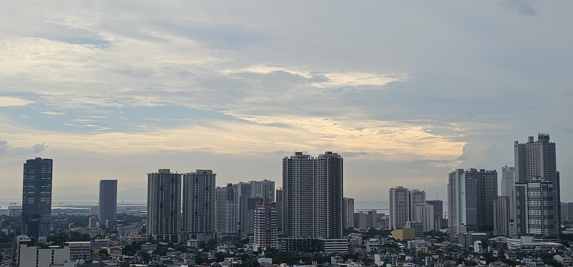 A wide-angle, eye-level, outdoor, daytime, establishing shot captures a sprawling urban cityscape dominated by numerous high-rise buildings, likely condominiums or office towers, situated in Makati, Philippines. The sky is partially overcast with dynamic cloud formations, some of which are illuminated by soft, warm light, suggesting either dawn or dusk. In the foreground and midground, a dense cluster of buildings of varying heights and architectural styles creates a sense of depth and scale. Distinctive tall, slender towers with reflective glass facades are visible, as are blockier structures with visible balconies or modular window designs. Some buildings appear to be under construction, with exposed framework and scaffolding. In the background, a body of water, possibly Manila Bay, is discernible, with faint outlines of distant landmasses or islands on the horizon. The overall impression is that of a bustling, modern metropolis viewed from an elevated vantage point. There are no people or discernible actions within the frame.