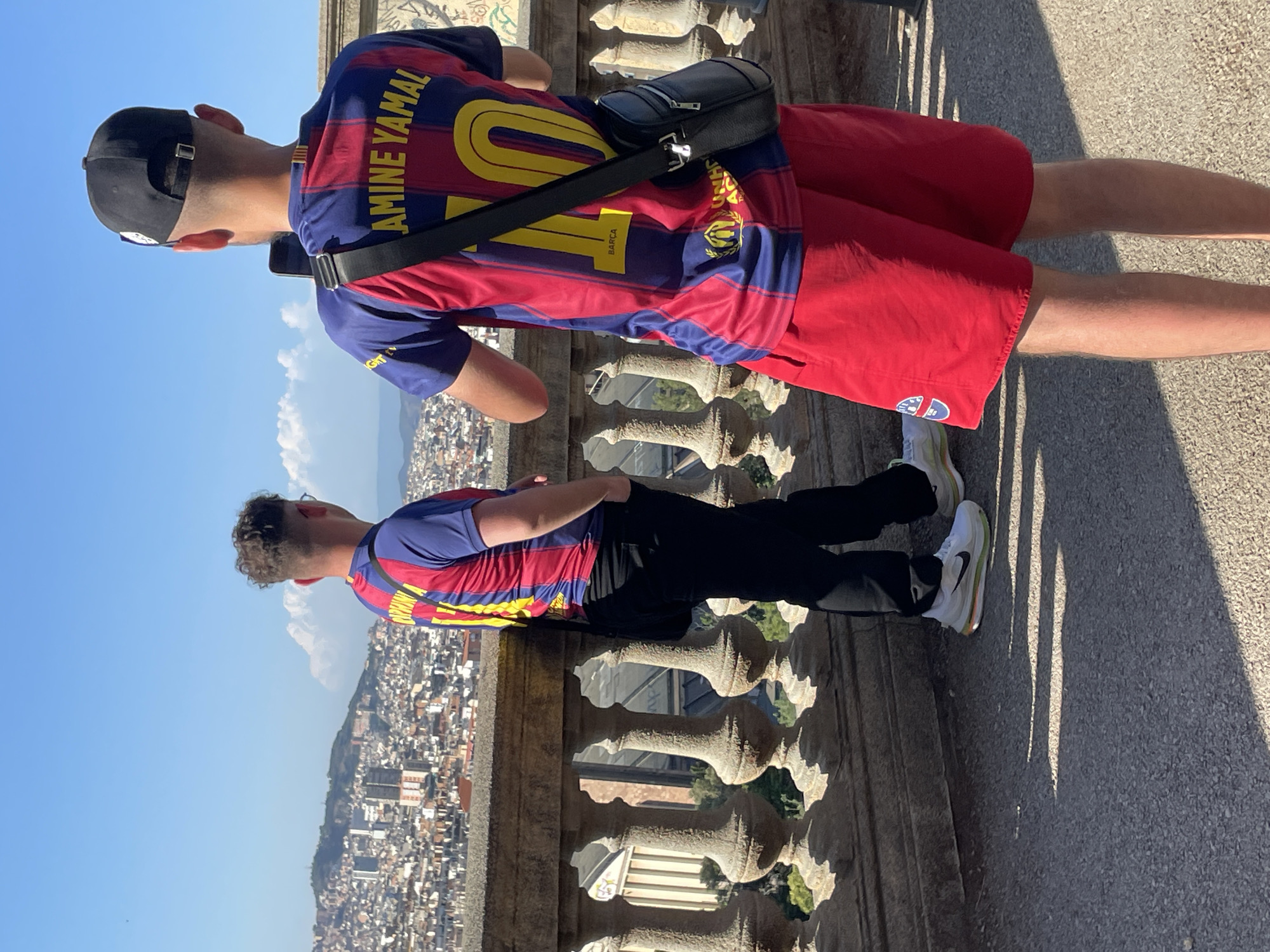Two young men are standing on a paved overlook, taking in a panoramic view of Barcelona, Spain. The city sprawls below, a dense urban landscape of white and terracotta rooftops, punctuated by taller buildings. In the foreground, a decorative stone balustrade with ornate balusters lines the edge of the overlook. The sky above is a clear, bright blue with a few wispy clouds.
The man on the left, facing away from the camera, is wearing a blue and red striped Barcelona football jersey with the number "10" on the back and "Gavi" across the nameplate. He is also wearing black pants and white sneakers. The man on the right, also facing away, is wearing a similar blue and red striped Barcelona football jersey with the name "AMINE YAMAL" and the number "10" on the back. He has on red shorts and is holding a smartphone, appearing to take a photo or video of the cityscape. A black crossbody bag is slung over his shoulder. The sun casts sharp shadows on the ground.