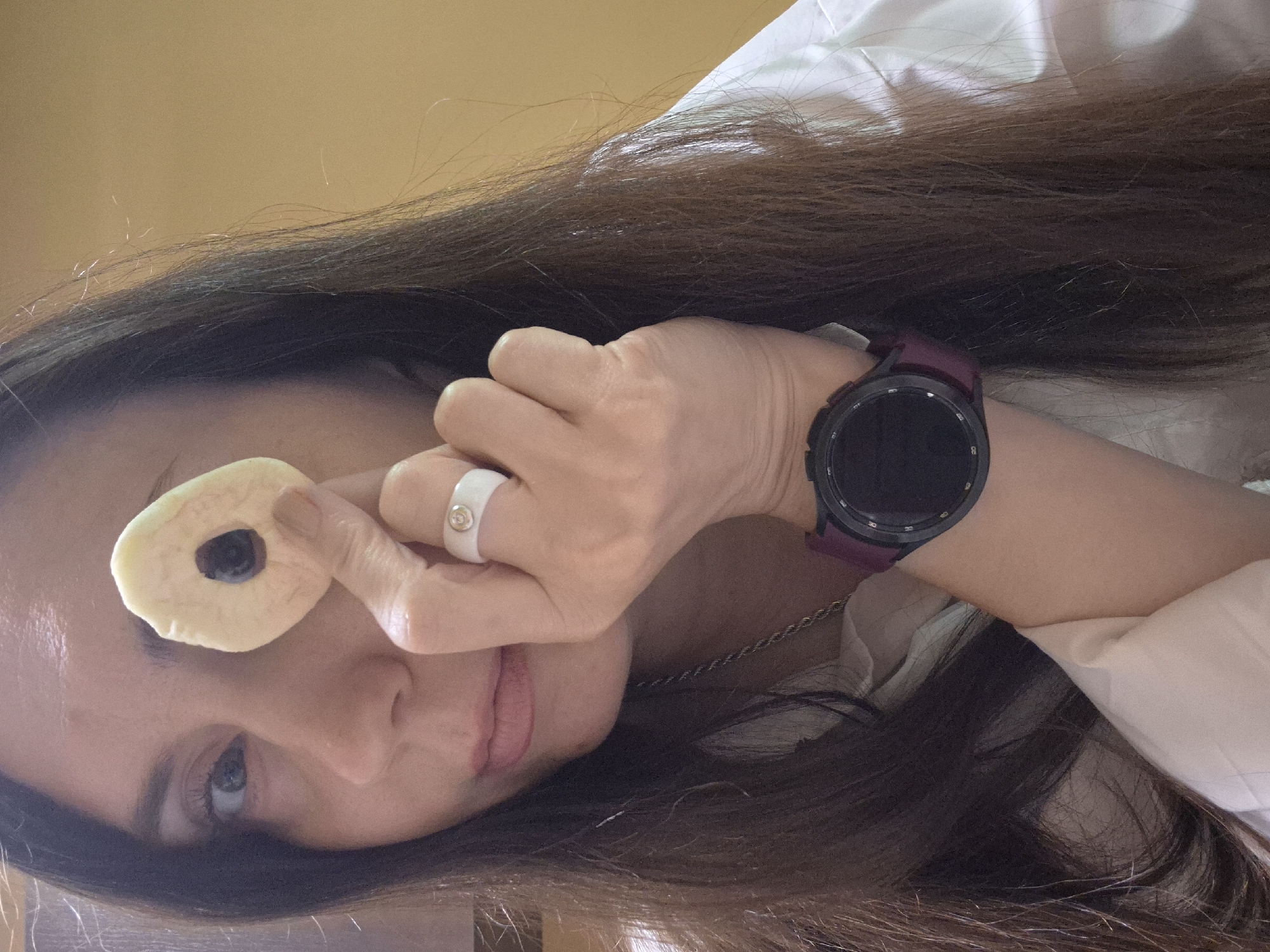 A woman is depicted in a close-up, sideways portrait. She is holding a donut-shaped object with a dark center in front of her right eye, as if looking through it. She wears a white ring on her right index finger and a smartwatch with a dark face and burgundy band on her left wrist. A delicate chain necklace is visible around her neck. Her long, brown hair is scattered around her head and shoulders. She has light skin, and the visible part of her face includes her eye, nose, and mouth. She is wearing a white shirt. The background is a solid, muted yellow color. The location is Unknown, Unknown.