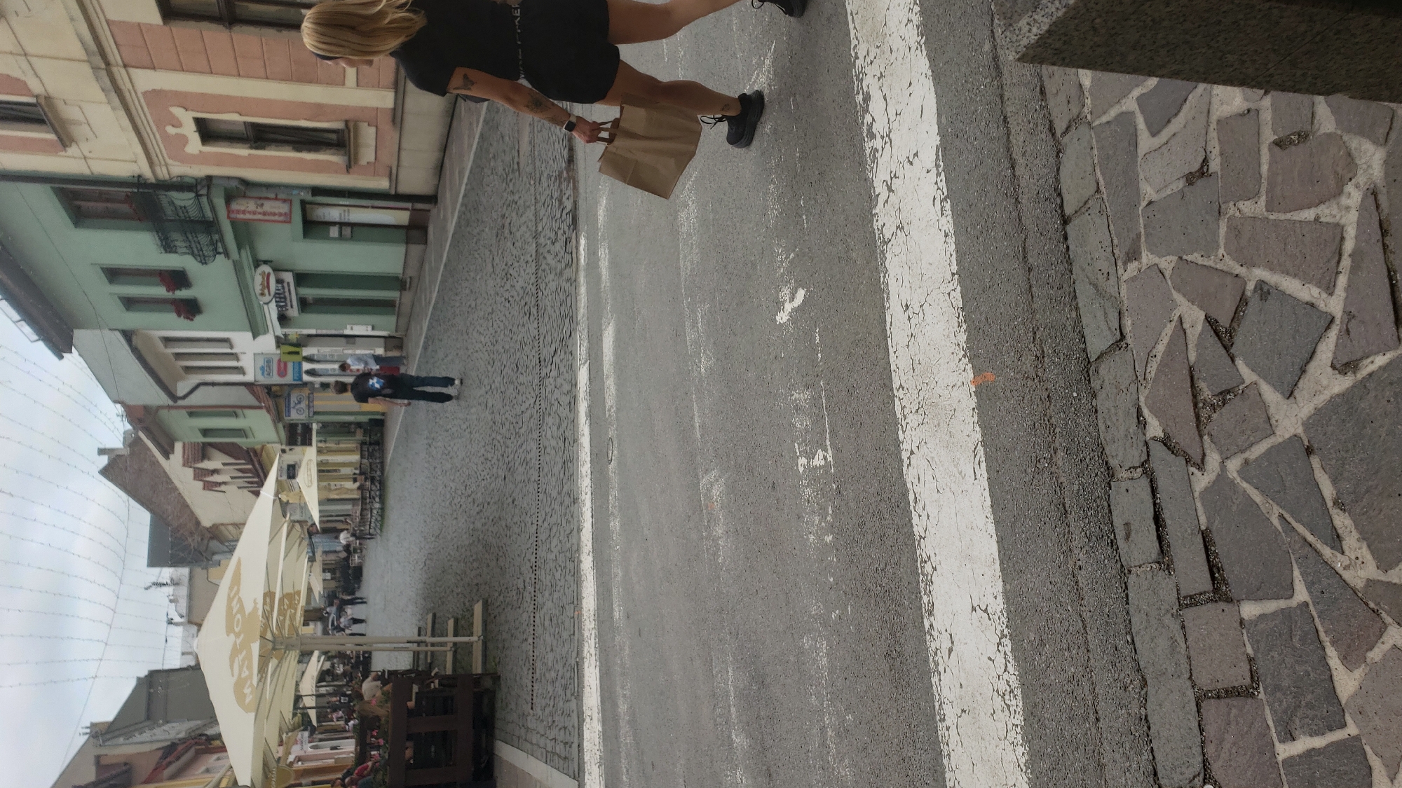 The image captures a street scene in Prešov, Slovakia, likely on a cobblestone street lined with buildings and outdoor seating areas. In the foreground, a woman walks across a pedestrian crossing, carrying a brown paper bag. She is wearing black shorts and a black t-shirt. Her legs and torso are visible from the knees up. The street is paved with asphalt and has white pedestrian crossing markings. Behind the woman, a young person in dark clothing walks away from the camera. Further in the background, outdoor umbrellas shade tables and chairs. The buildings on either side of the street exhibit varied architectural styles and colors, with some featuring balconies and shopfronts. String lights are visible across the street overhead. The sky is overcast.