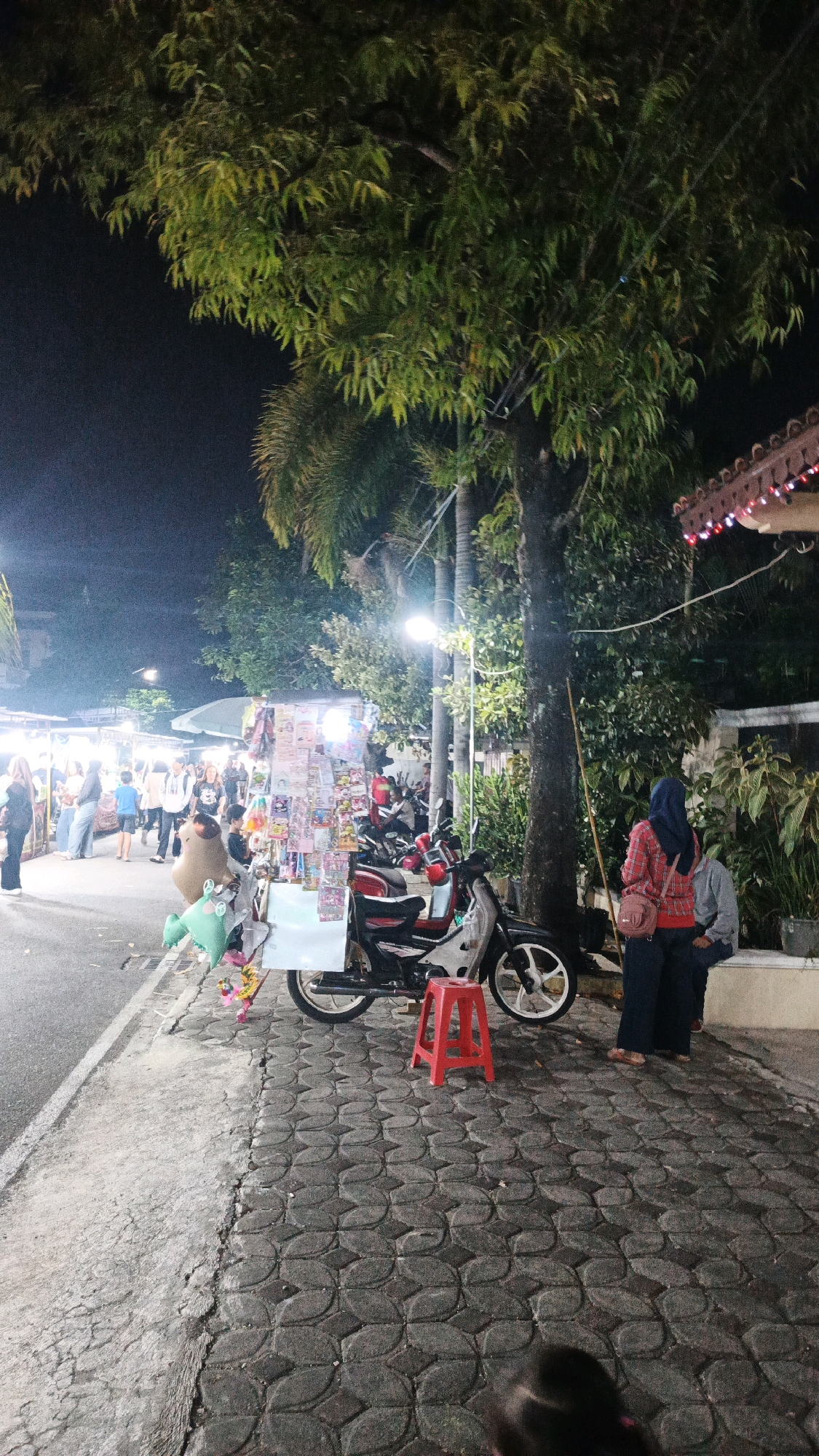 This image depicts a nighttime street scene in Surakarta, Indonesia, likely at a local market or a busy roadside area. A street vendor's cart, laden with various items including stuffed toys, is parked alongside the road. A motorcycle is parked behind the cart, with a red stool in front of it. Two individuals, one wearing a red and black plaid shirt and a blue hijab, and another person partially visible, stand near the cart. In the background, several other people are seen walking along the street, and stalls with merchandise are visible under the glow of streetlights and artificial illumination. The ground is paved with interlocking bricks and concrete. Lush green trees and palm fronds overhang the scene, illuminated by the lights.