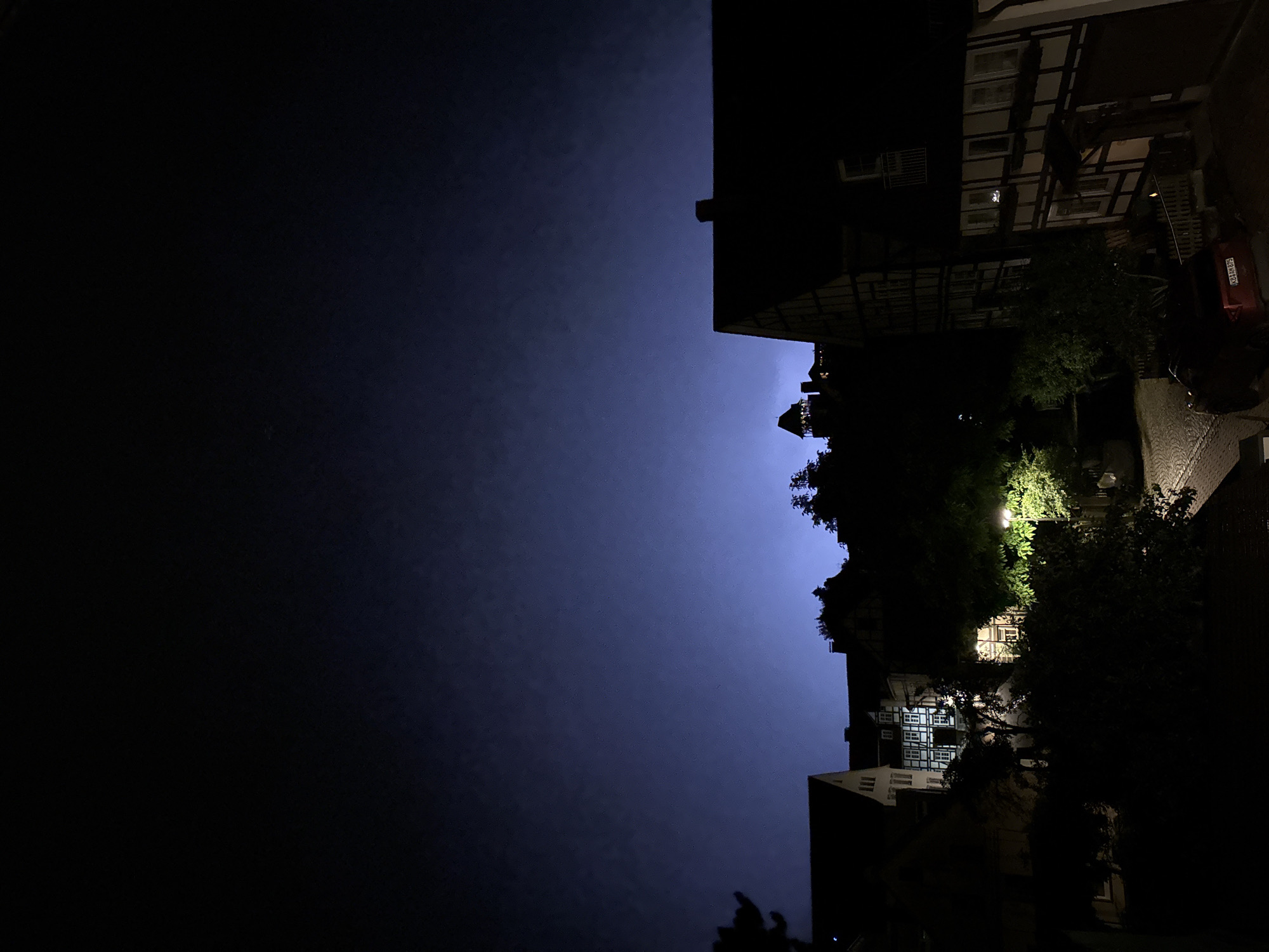 A night time, low-angle, wide shot captures a European town at dusk, with a prominent, dark blue sky. The sky has subtle cloud formations, and a faint, distant light emanates from the horizon, suggesting a storm or distant city lights. In the foreground and midground, silhouetted buildings with traditional architecture, including half-timbered structures, are visible. Trees and foliage obscure parts of the scene. Several buildings have illuminated windows, indicating occupancy. A streetlamp shines brightly, illuminating a cobblestone street and part of a red car parked on the right side of the frame. The car has a license plate that reads "KB AM 875." In the far distance, a tower or castle structure is visible on a hilltop, also faintly illuminated. The overall mood is serene yet ominous due to the twilight and impending weather.