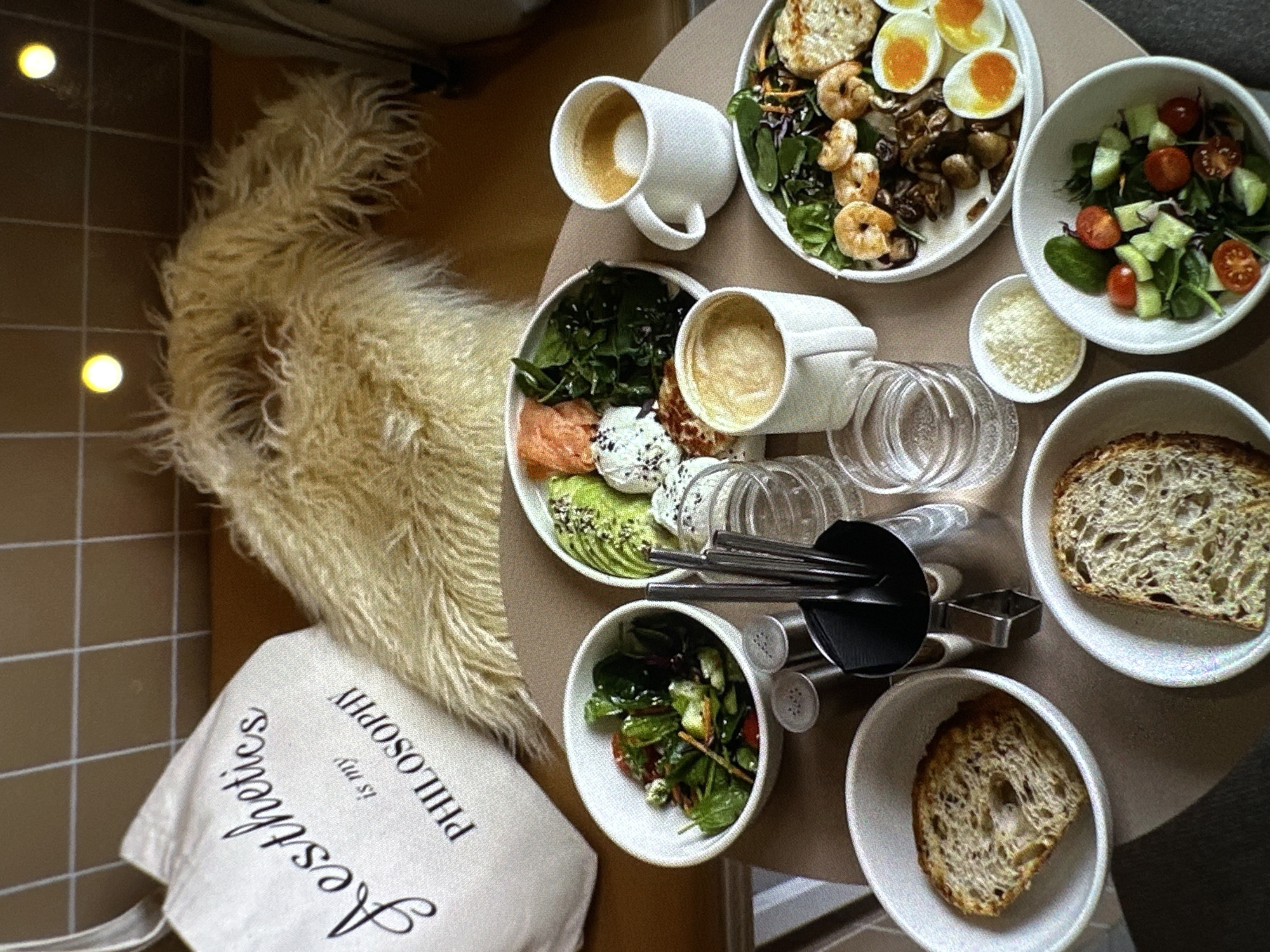The image shows a tabletop scene with a variety of food and drink items, as well as a few other objects. There are two bowls of salad, one with tomatoes and cucumbers, and another with leafy greens. One of the bowls contains what appears to be a salmon and avocado salad with other unidentified elements. There are also two slices of bread in bowls. One plate includes a mix of grilled mushrooms, shrimp and boiled eggs. There are two white ceramic coffee mugs, each containing a beverage that looks like coffee or latte. A small bowl contains what appears to be grated cheese. There are clear drinking glasses and cutlery present on the table as well.
A furry dog lies adjacent to a white tote bag with the phrase "Aesthetics is my PHILOSOPHY" printed on it. The tabletop is covered with a neutral-colored surface. The image is taken from an overhead perspective.
There is no definitive information in the image to determine the location in Yerevan, Armenia.