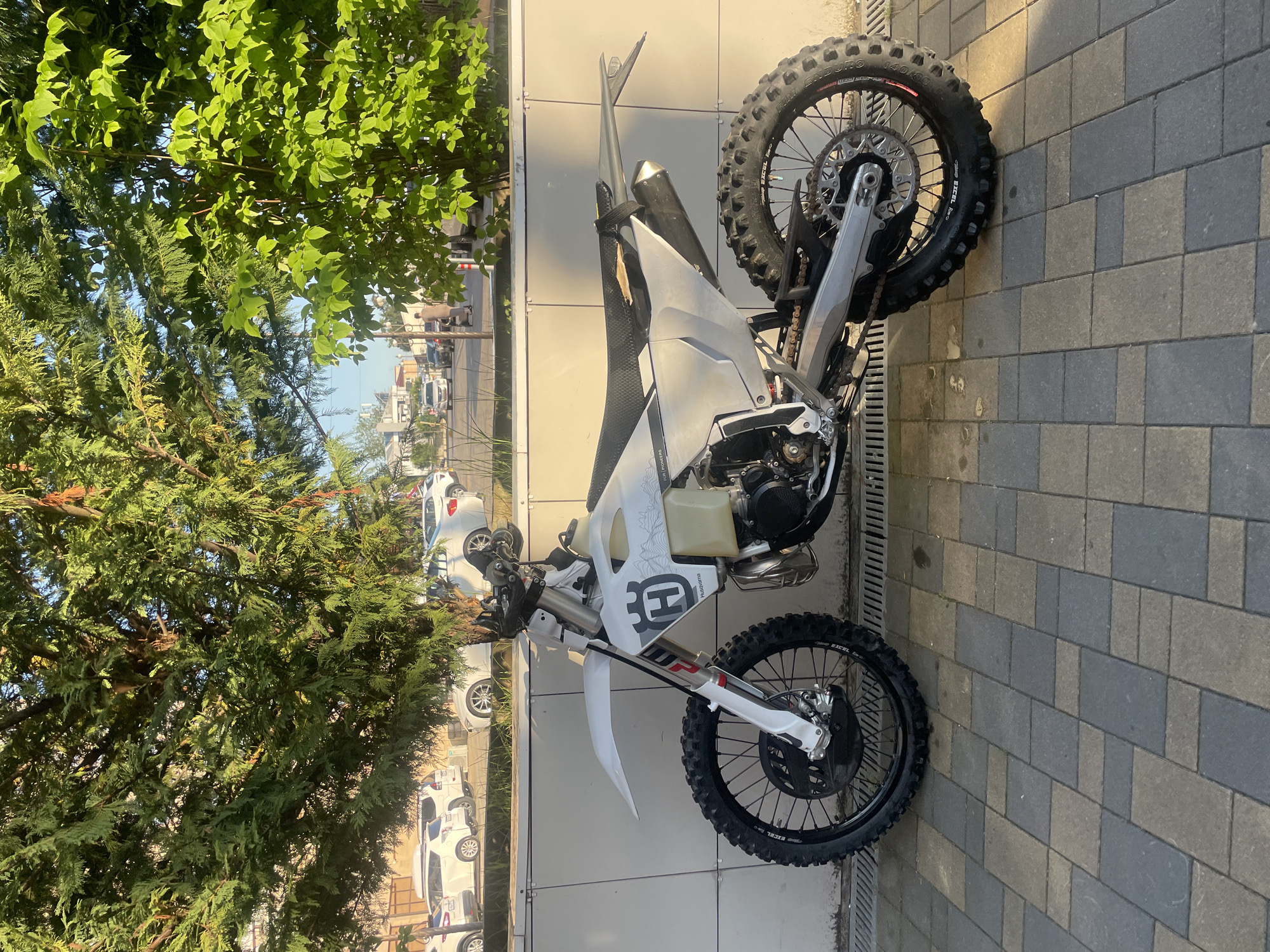 A white Husqvarna dirt bike is prominently parked in profile on a patterned block paving sidewalk in Sochi, Russia. The motorcycle faces left, displaying its white body panels, black knobby tires on black Excel rims, and visible suspension components, including a white front fender and red/silver forks. The fuel tank features the Husqvarna logo and an "H" emblem, along with a subtle topographical pattern. The black seat appears worn with visible damage. A black exhaust muffler is positioned on the right side of the bike.
Behind the motorcycle is a light-colored building wall constructed from horizontal panels. A metal drainage grate runs along the base of the wall and sidewalk.
The background features lush green foliage from two distinct trees: a dense evergreen with fine, scale-like leaves on the left, and a deciduous tree with broader, lighter green leaves on the right. Beyond the trees, a paved street is visible with several white and light-colored cars parked or driving. In the middle-ground, a person dressed in light brown clothing is seen walking away from the camera, pushing a dark object. The sky above is clear and light blue. The scene is illuminated by strong, direct sunlight, casting shadows on the pavement and wall, suggesting a clear day, likely morning or late afternoon.