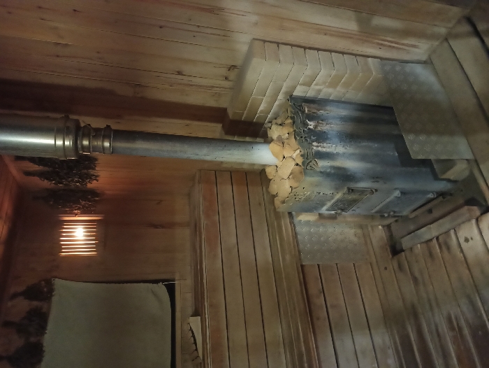 The image displays the interior of a banya or sauna in Nizhneye Zadolgoye, Russia. The room features light-colored wooden plank walls, floor, and ceiling. A metallic, dark-colored wood-burning stove stands on the right, filled with light-colored stones on its upper surface. A stainless steel flue pipe extends vertically from the stove. Tiered wooden benches are present along the left wall. A slatted light fixture on the left wall illuminates the space with a warm glow. Several bundles of dried branches (veniks) hang from the wooden walls. A light-colored cloth or towel is draped over the uppermost bench on the left. Diamond-plate metal panels serve as protective barriers behind and in front of the stove, complemented by a brick-like structure on the adjacent right wall. No individuals are present in the scene.