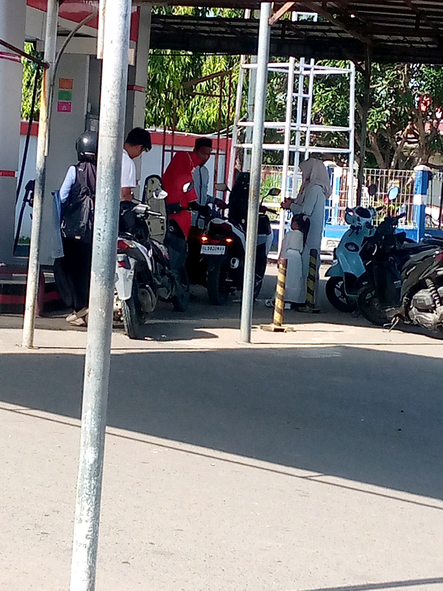 At a gas station in Keudebayu, Indonesia, several individuals are gathered around motorcycles. One person, wearing a red jacket and a black head covering, is interacting with a motorcycle. Another individual, in a light blue hijab and dress, is standing near a child who is also dressed in light blue. A person with a black helmet and backpack stands near a gas pump. In the background, trees and a white fence are visible. The scene is lit by daylight.