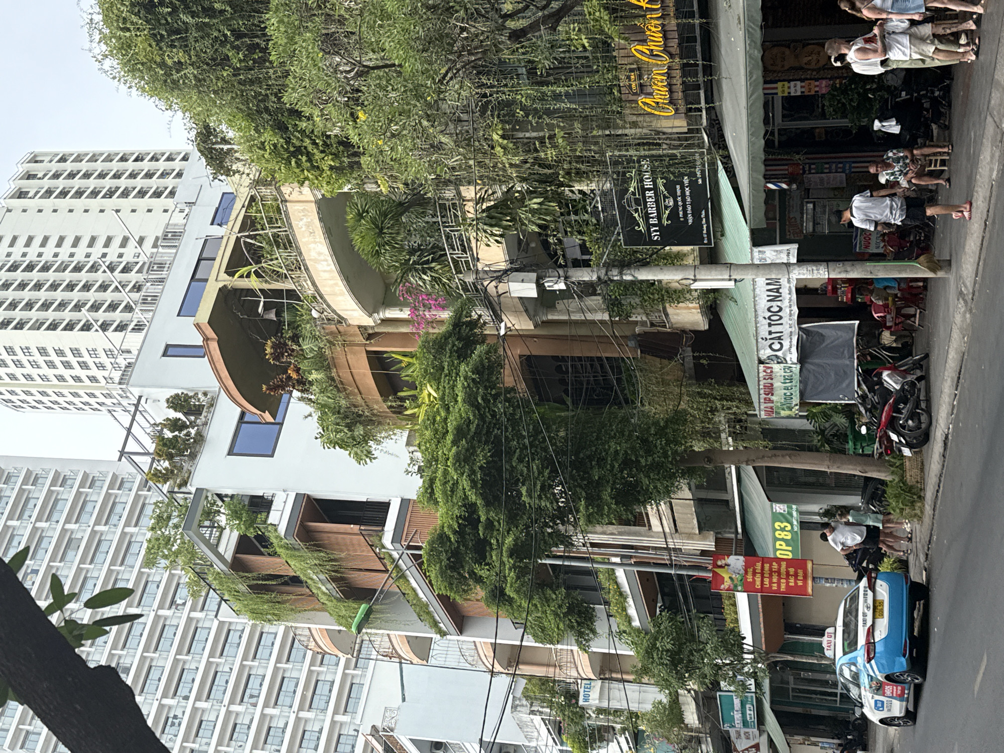 Here's a concise description of the scene:
The image depicts a street scene in Nha Trang Ward, Vietnam. A tall, modern residential building stands in the background on the left. Closer to the viewer, there are multi-story buildings with balconies adorned with greenery. Overhead power lines crisscross the scene. On the right side of the street, there's a barber shop ("Syy Barber House") with customers waiting outside. Nearby, there's a blue-and-white taxi and other parked motorbikes. Street signs and advertisements are visible. A sign above the barber shop is written in Vietnamese and an unidentifiable word. The general environment suggests a mix of residential and commercial activity.
