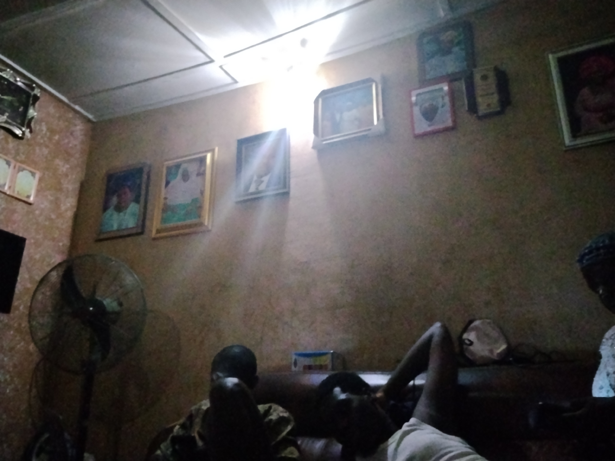 The photograph appears to be taken indoors in a residential setting in Shangisha, Nigeria. The room is dimly lit, with light streaming from an overhead fixture. The walls are covered in patterned wallpaper and decorated with several framed pictures. An electric fan is visible in the left side of the frame. Three people are visible: one person is partially obscured in the lower left corner, one is lying down in the center, and another person is partially seen in the lower right corner. The individuals are resting on what appears to be a sofa. Additional objects are present on a surface behind them.
