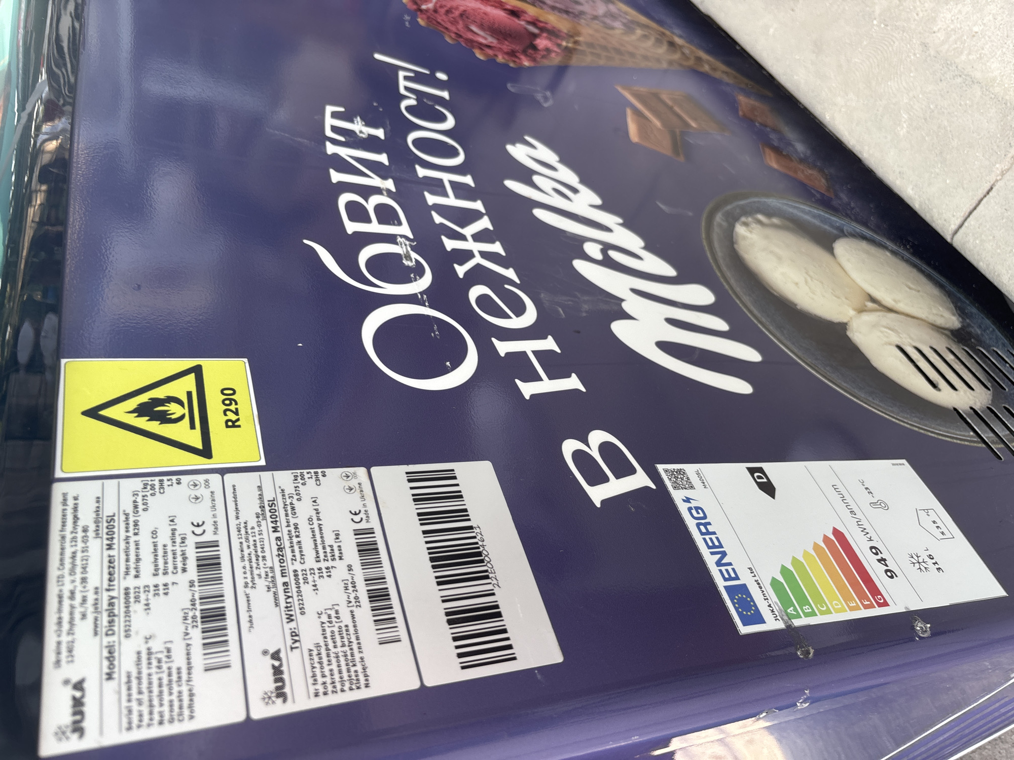 The image displays a JUKA brand commercial display freezer, model M400SL, located in the city of Kostievo, Bulgaria. The freezer unit features a dark blue exterior adorned with several informational labels and a prominent advertisement.
Two primary product labels are affixed to the upper left side of the freezer. The top label, in English, identifies the manufacturer as Ukraine "Juka-Invest" LTD and lists specifications: serial number 05222004089 ("Hermetically sealed"), production year 2022, R290 refrigerant (0.075 kg), temperature range -14 to -23 °C, net volume 316 L, gross volume 416 L, climate class 7, current rating 1.5 A, voltage 220-240~/50 V/Hz, and weight 60 kg. A second label, in Polish, is located directly below the English one, presenting identical technical data. Both labels bear a CE mark and state "Made in Ukraine." A barcode, partially visible below the Polish label, contains the number "22E0004621".
To the right of the product labels, a yellow triangular warning symbol with a flame icon clearly states "R290", denoting the refrigerant type.
An EU ENERG energy efficiency label is present on the lower left of the freezer, indicating the JUKA-Invest Ltd M400SL model's energy class as D, with an annual consumption of 949 kWh/annum. This label also specifies a storage volume of 316 L and a freezing temperature of -23 °C.
The majority of the freezer's front surface is covered by an advertisement featuring white Cyrillic text: "ОБВИТ НЕЖНОСТ!" and the stylized brand name "Milka". The graphic depicts three scoops of white ice cream or frozen dessert in a metallic bowl with a serving utensil on the left. On the right, part of an ice cream cone filled with pink-purple ice cream is visible, alongside scattered rectangular chocolate pieces in the background.
The freezer is situated on a light-colored tiled floor with discernible grout lines. No individuals are visible in the scene.