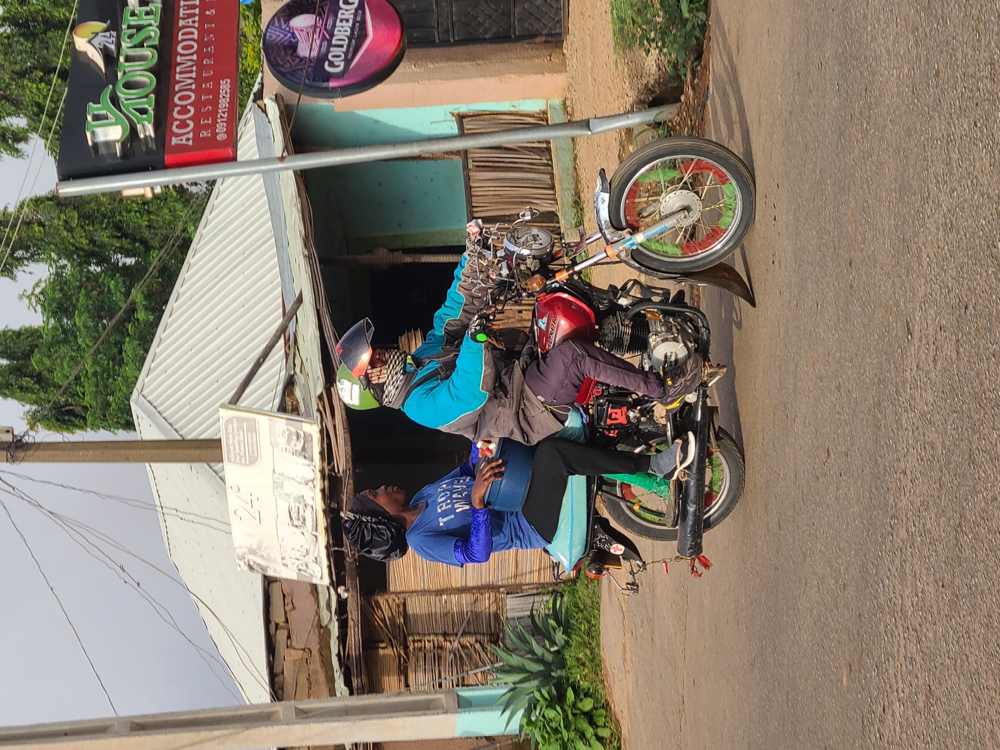 The image shows two people on a motorcycle in an urban area. The driver, wearing a blue jacket and helmet, is operating the motorcycle. A passenger in a blue shirt sits behind, carrying what appears to be a blue container. The background includes buildings with signage indicating "24 House Accommodations Restaurant and Bar" and "Goldberg." The environment seems to be a roadside in the city of Jos, Nigeria.