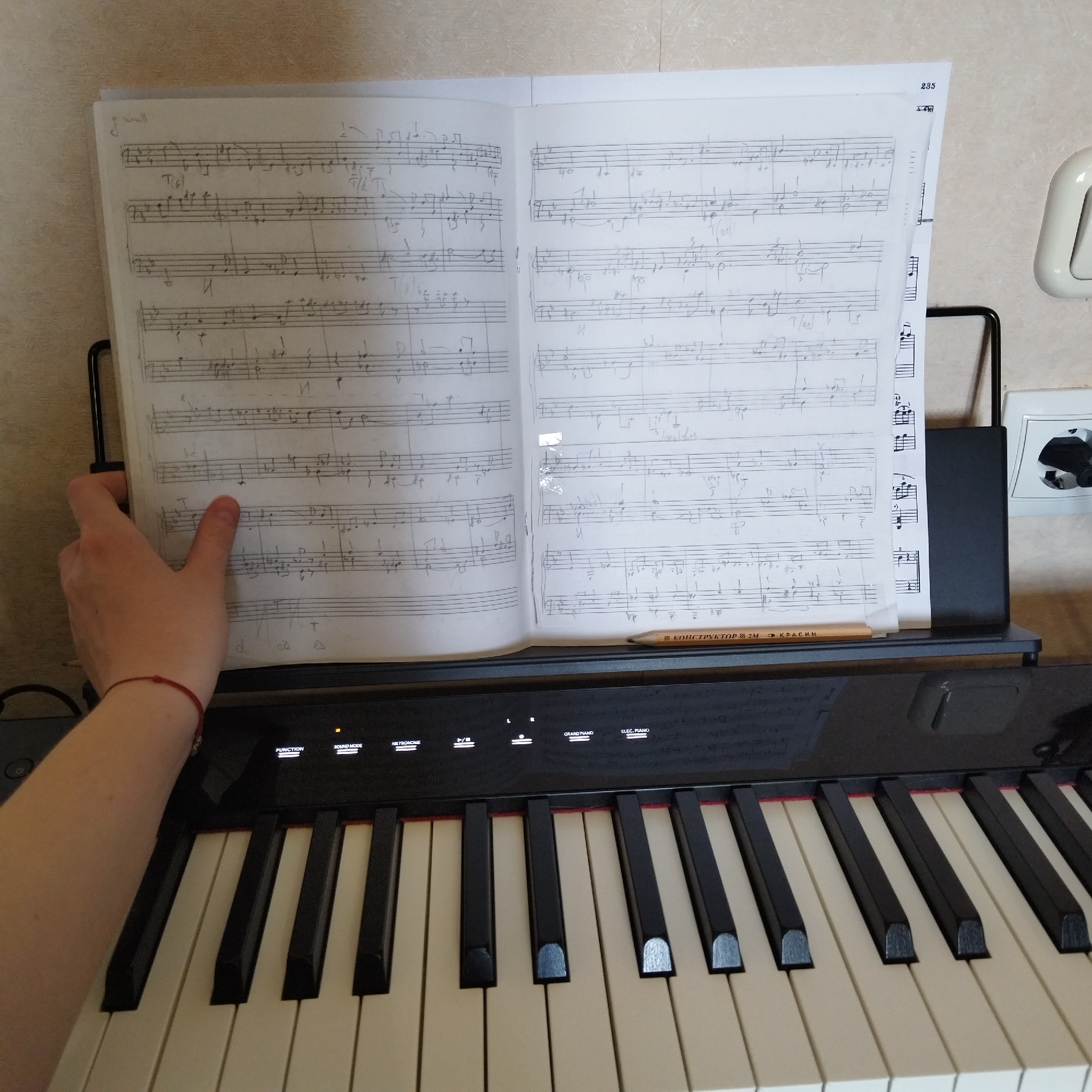 A slightly overhead, medium shot captures a person's left hand and forearm reaching towards an open music book on a digital piano. The scene is indoors, with a textured, light-colored wall visible in the background.
The primary subject is a digital piano with a black, reflective control panel and standard white and black keys. Above the keys, on a black music stand, an open book displays two pages of handwritten or photocopied musical notation. The left hand of the person is positioned over the left page of the music book, appearing to either hold it open or prepare to turn the page. A thin red string is tied around the person's left wrist. A pencil rests on the piano's control panel, below the right page of the music. To the right of the music stand, a white wall socket with a black plug is visible.
Notable details include the intricate musical symbols, notes, and staves on the sheet music. A page number "235" is visible in the top right corner of the right page. The piano's control panel shows illuminated text labels: "FUNCTION", "SOUND MODE", "METRONOME", "GRAND PIANO", and "ELEC. PIANO", along with " /4" which might indicate a tempo or time signature setting. The pencil has Cyrillic text, "КОНСТРУКТОР 2М КРАСИН", printed on it. The overall setting suggests a music practice or study environment.
