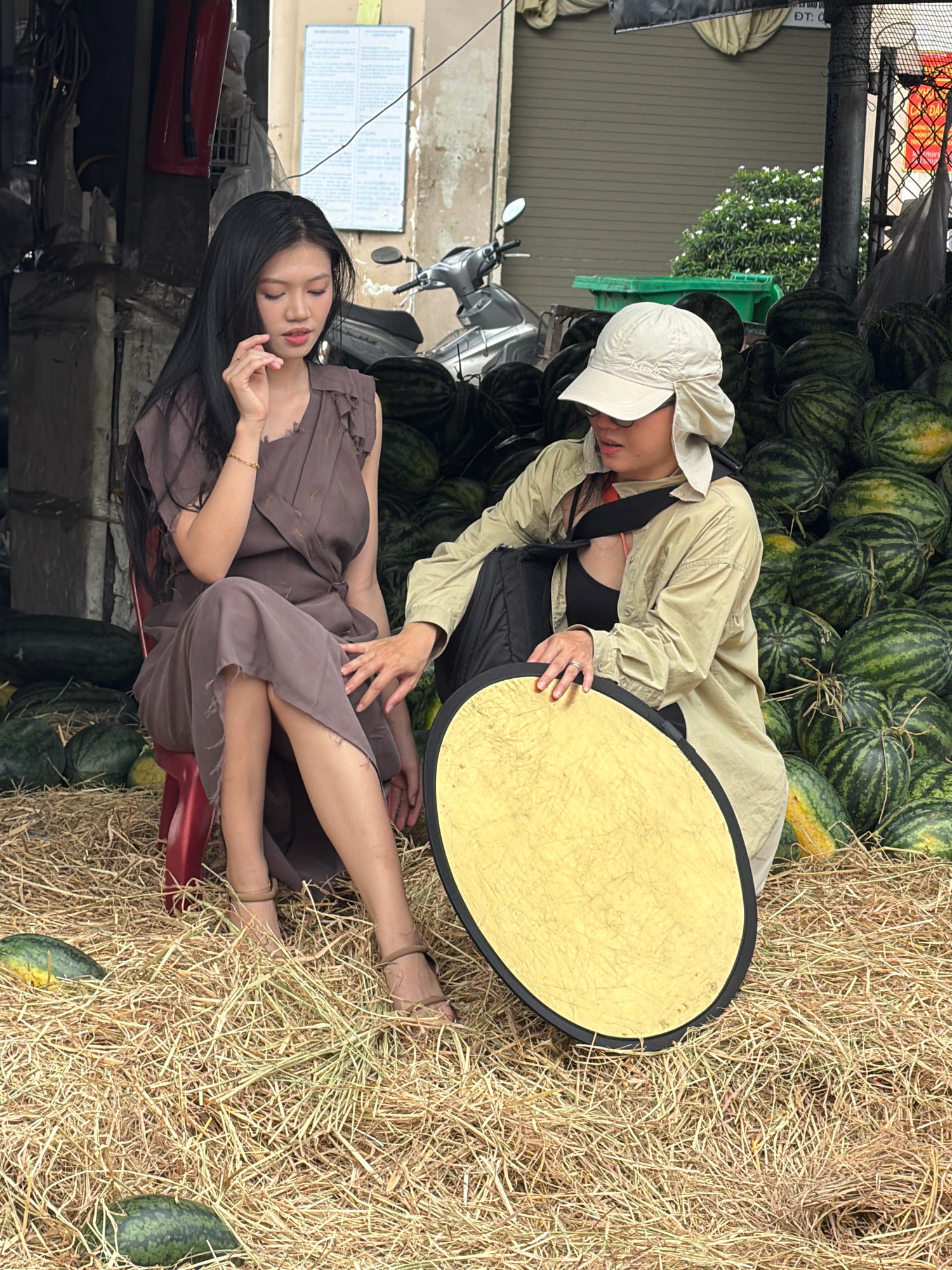 The image depicts an outdoor scene in Nha Trang, Vietnam, likely at a marketplace, featuring two individuals amidst a large pile of watermelons.
On the left, a young woman with long, dark hair sits on a small red plastic stool. She is wearing a loose-fitting, dusty brown dress with distressed edges and has a delicate posture, with one hand near her face and the other resting on her lap. She has a bracelet on her left wrist.
To her right, another person, wearing a light-colored hat with ear flaps, a light-colored long-sleeved shirt, and a black tank top underneath, is kneeling and holding a round, golden reflective disc with a black rim. Their hands are visible, with the person on the right resting their hand near the disc and the woman on the left extending her hand towards the disc.
The ground is covered in dry hay, with several watermelons scattered around. The background shows a stack of what appear to be concrete blocks or large crates, and further back, a motorcycle is visible. A green trash bin and more watermelons are piled high behind the individuals. The overall environment suggests a rustic, possibly commercial setting.