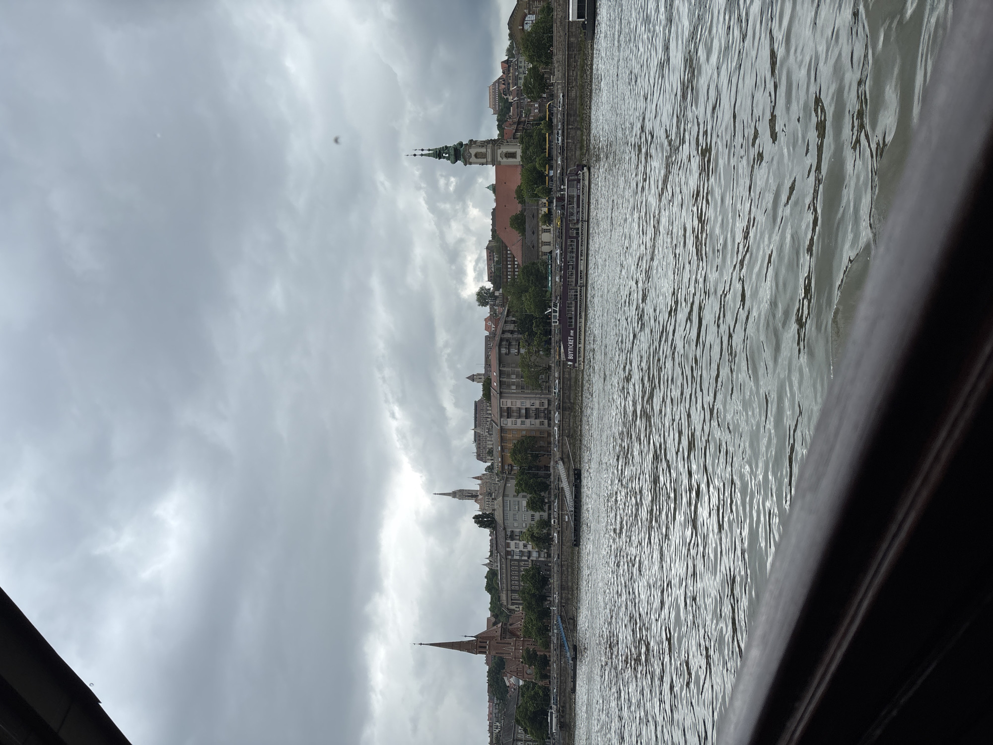 The image depicts a cityscape in Budapest, Hungary, captured from a boat on the Danube River. The buildings along the riverbank include several prominent spires and towers, suggesting the presence of churches and other historical structures. The sky is overcast with dense cloud cover. The Danube River is the most prominent physical feature and there are boats docked along the river. The perspective is from a low angle, likely from the deck of a boat, partially obstructed by a dark, possibly wooden, railing.