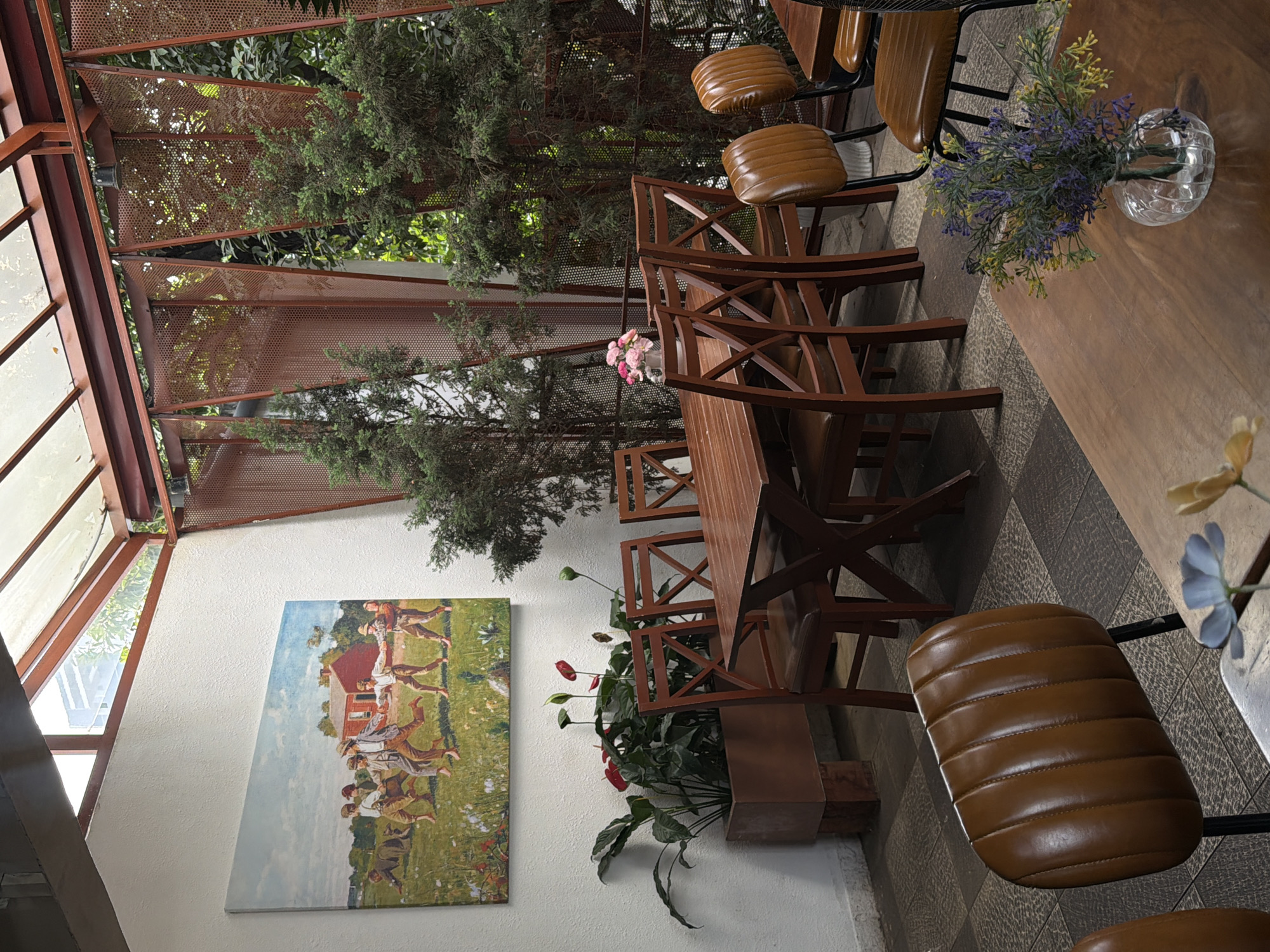 This media file presents a brightly lit, semi-open dining or cafe area, likely located in Đà Nẵng, Vietnam. The space blends industrial and natural aesthetics, characterized by its reddish-brown metal framework and lush greenery.
The scene features multiple wooden tables paired with matching wooden chairs, which are fitted with comfortable, ribbed brown leatherette cushions. The flooring is composed of patterned dark and light gray tiles. Natural light streams into the area, indicating it is daytime with clear weather.
Green trees and foliage are prominently visible beyond the perforated metal ceiling panels and through glass sections of the roof, creating an indoor-outdoor feel. Decorative plants are also present within the space; a vibrant plant with red, spade-shaped leaves is housed in a wooden planter against a white wall. Smaller glass vases with artificial flowers, one with pink blooms and another with purple and yellow ones, adorn the tabletops.
A notable feature is a large painting hung on the white wall. It depicts a pastoral landscape with a group of individuals, possibly children, dressed in historical attire, engaged in a circular dance or game in a grassy field. A red-roofed building is visible in the background of the artwork.
The overall atmosphere is calm and inviting, suggesting a comfortable place for relaxation or dining. No people are present in the immediate seating area, and no visible text is discernible anywhere in the image.